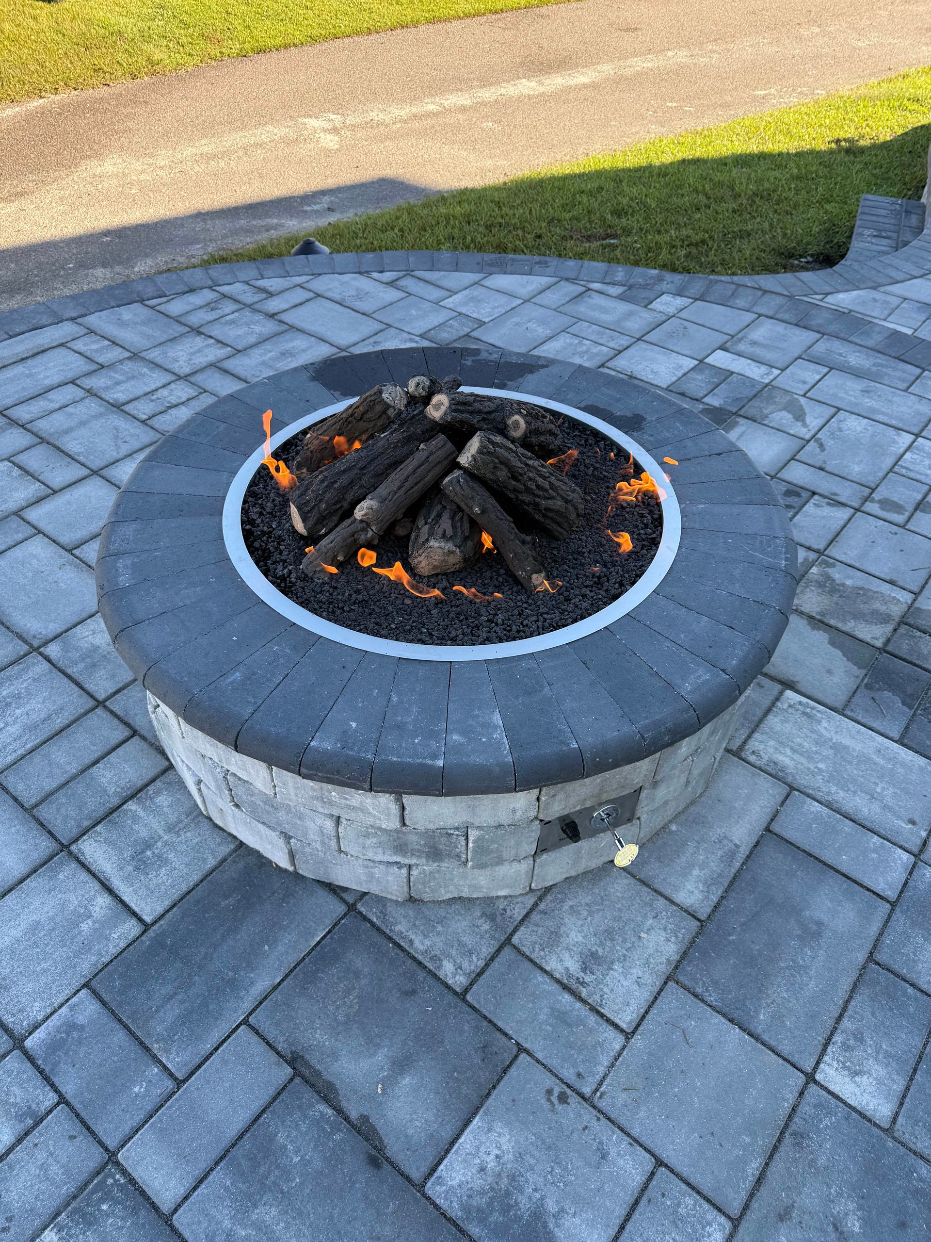 A circular stone fire pit with burning logs, on a paved patio.