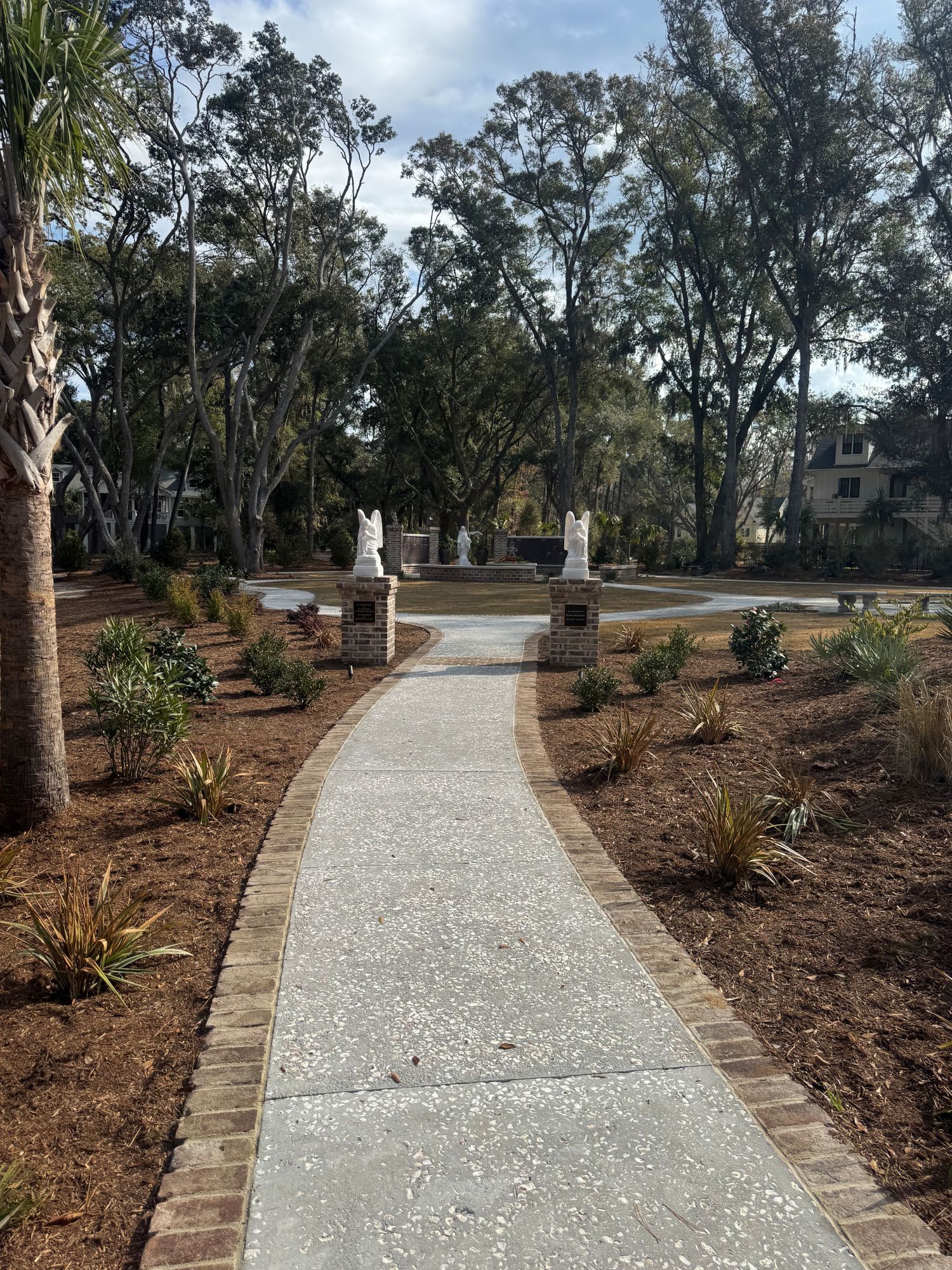 A brick-lined, concrete path leads to white sculptures in a garden setting with trees and brown mulch.