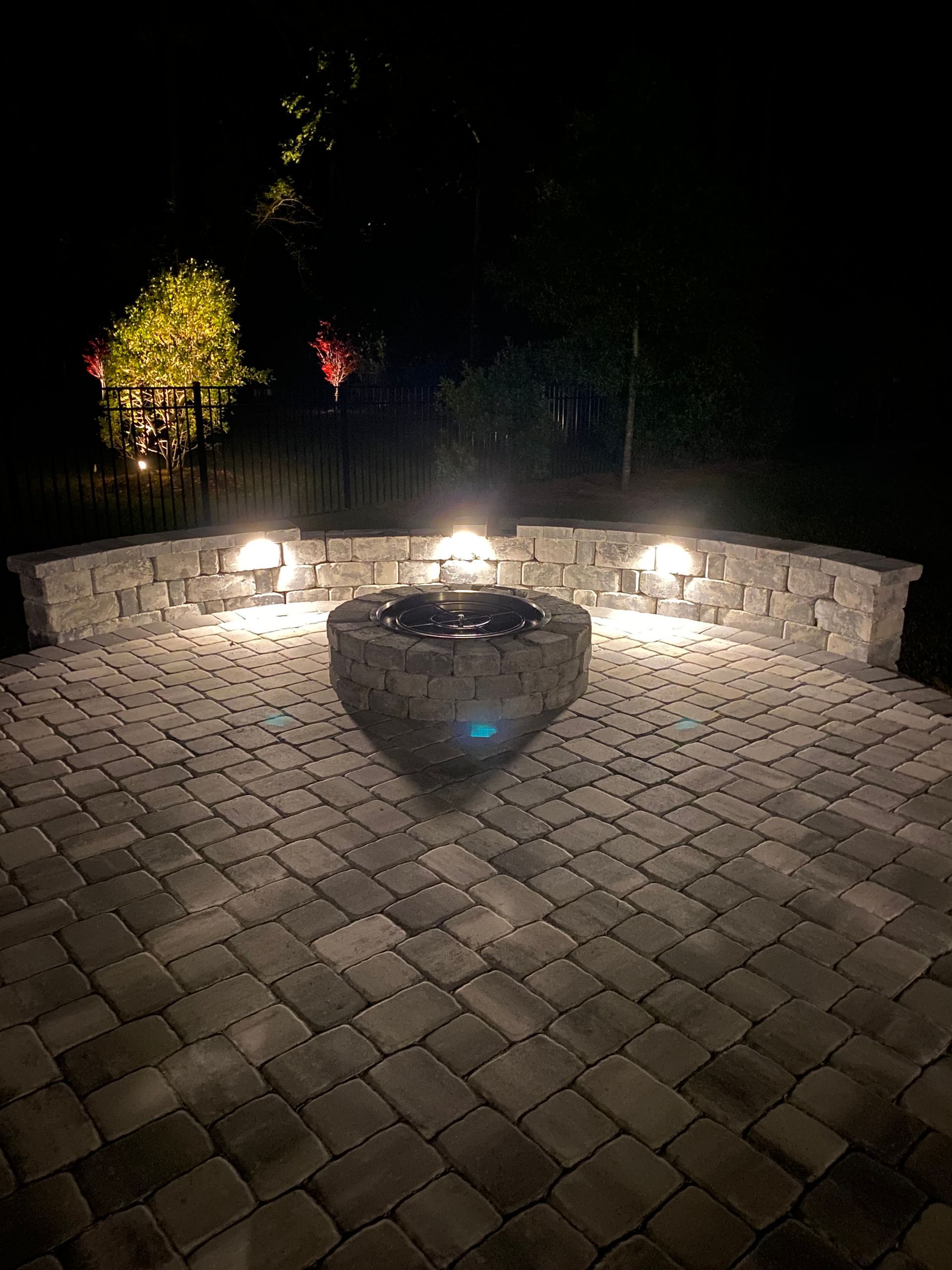 Nighttime patio with lit fire pit and stone wall. Brick paved floor. Three lights on the wall. Trees in the background.