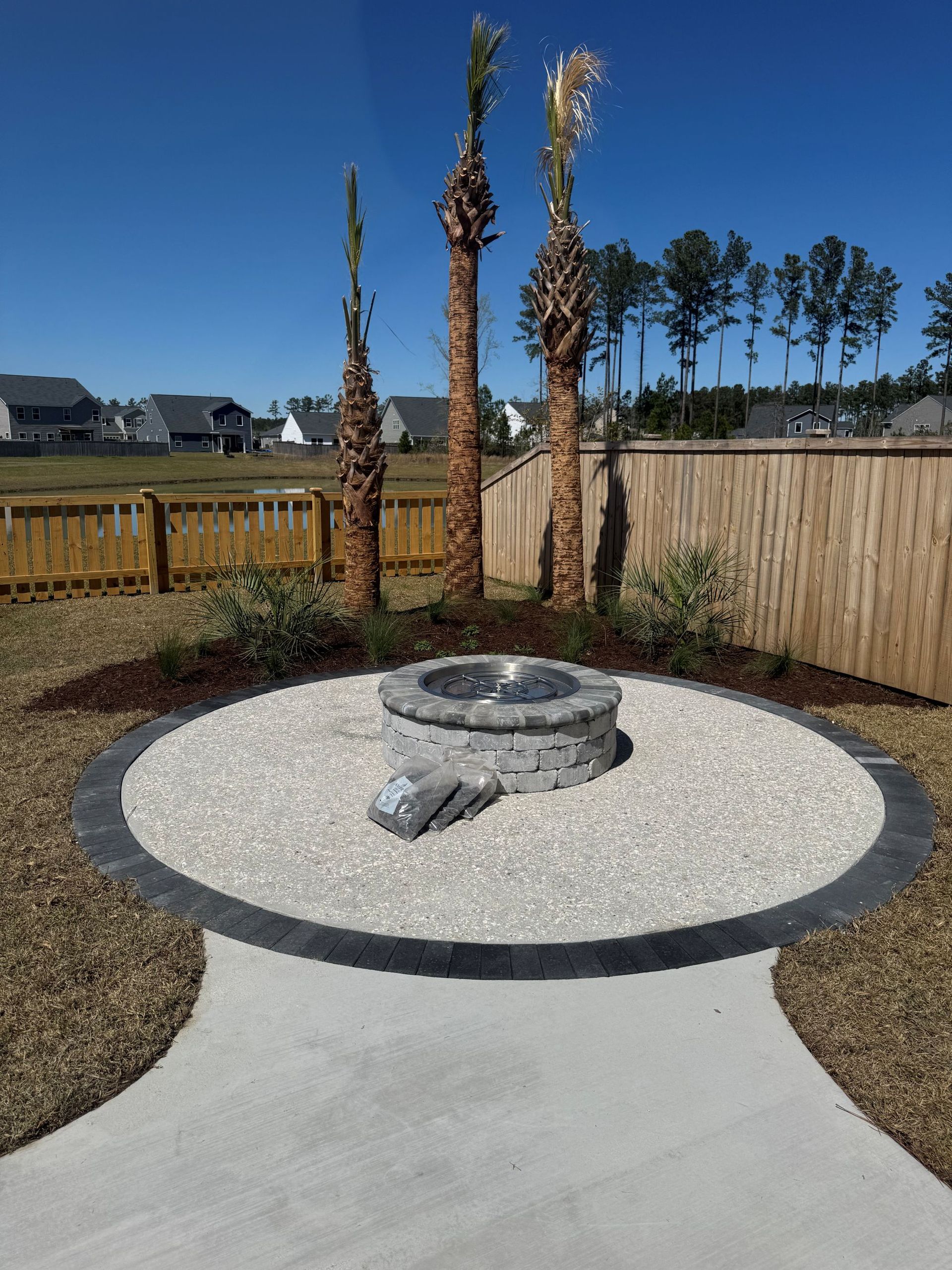 Fire pit on a stone patio with a concrete path. Palm trees and fence in the background under a blue sky.