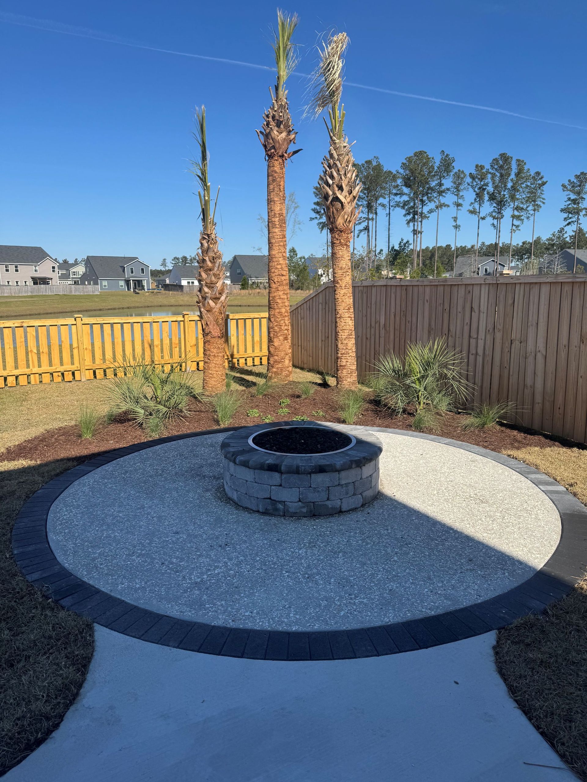 Backyard fire pit with stone surround and palm trees against a fence.
