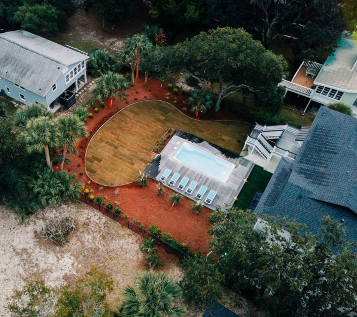 Aerial view of a backyard with a pool, landscaping, and adjacent houses. Brown grass, red mulch, blue pool.