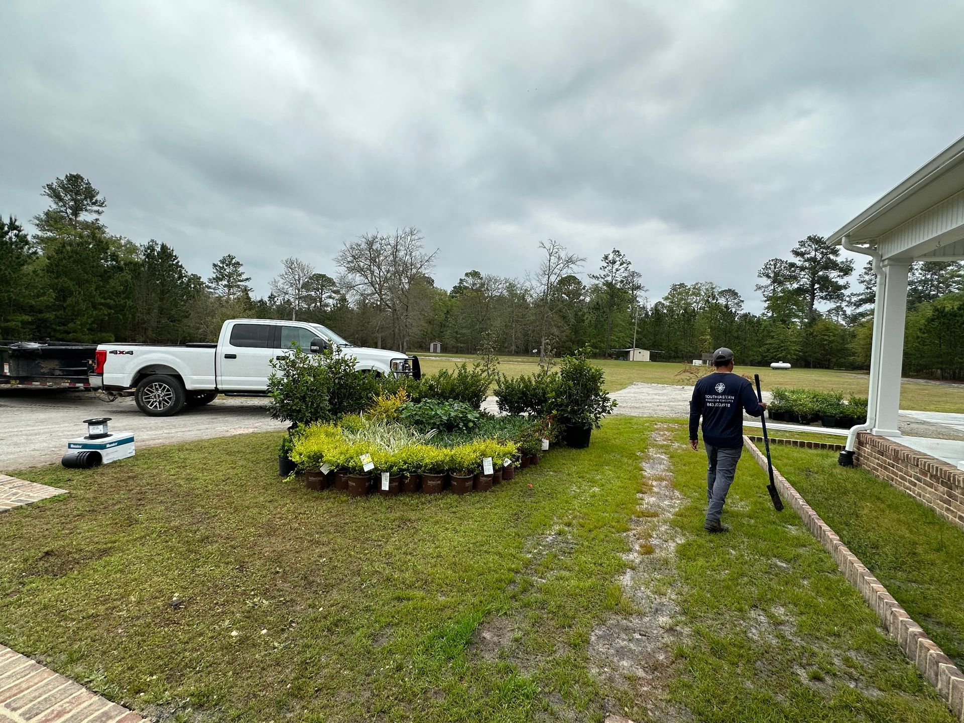 Man carries a shovel toward a house. A truck and trailer with plants are on the lawn. Overcast sky.