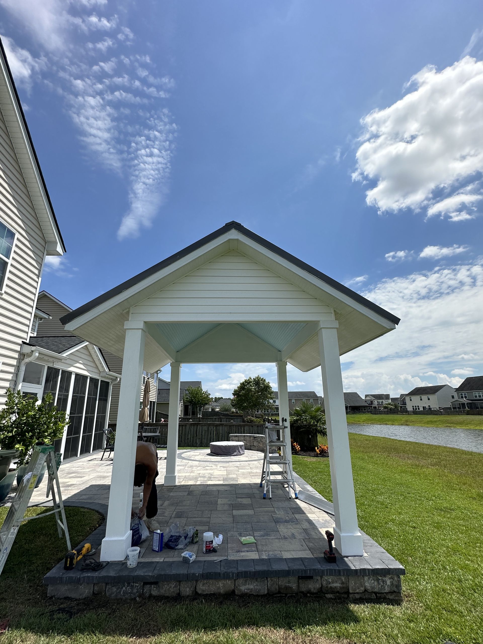 Gazebo on a patio, painted white with a dark gray roof, blue sky, grassy yard, and pond in the distance.