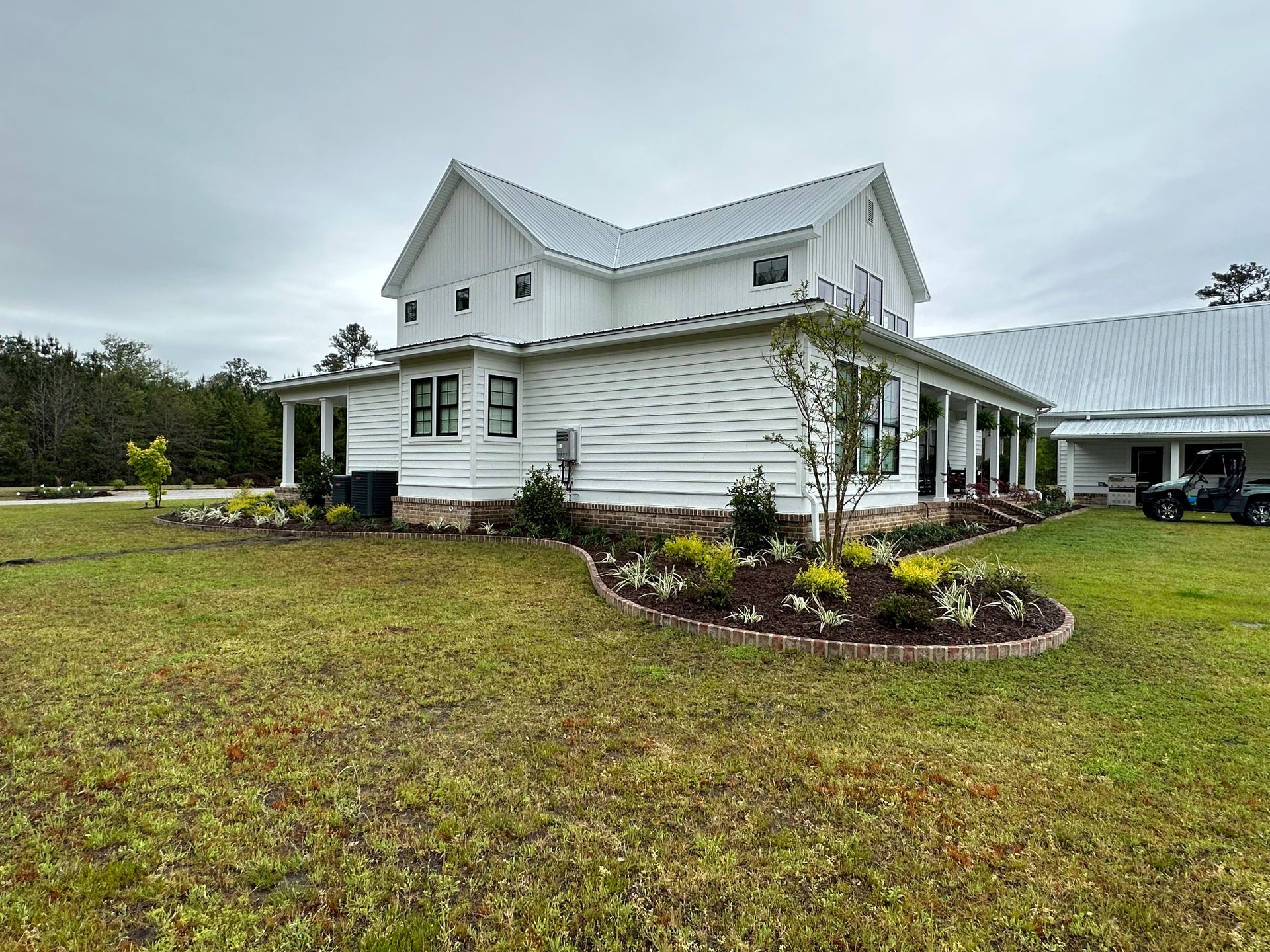 White farmhouse with metal roof and porch, flower beds in front yard under a cloudy sky.