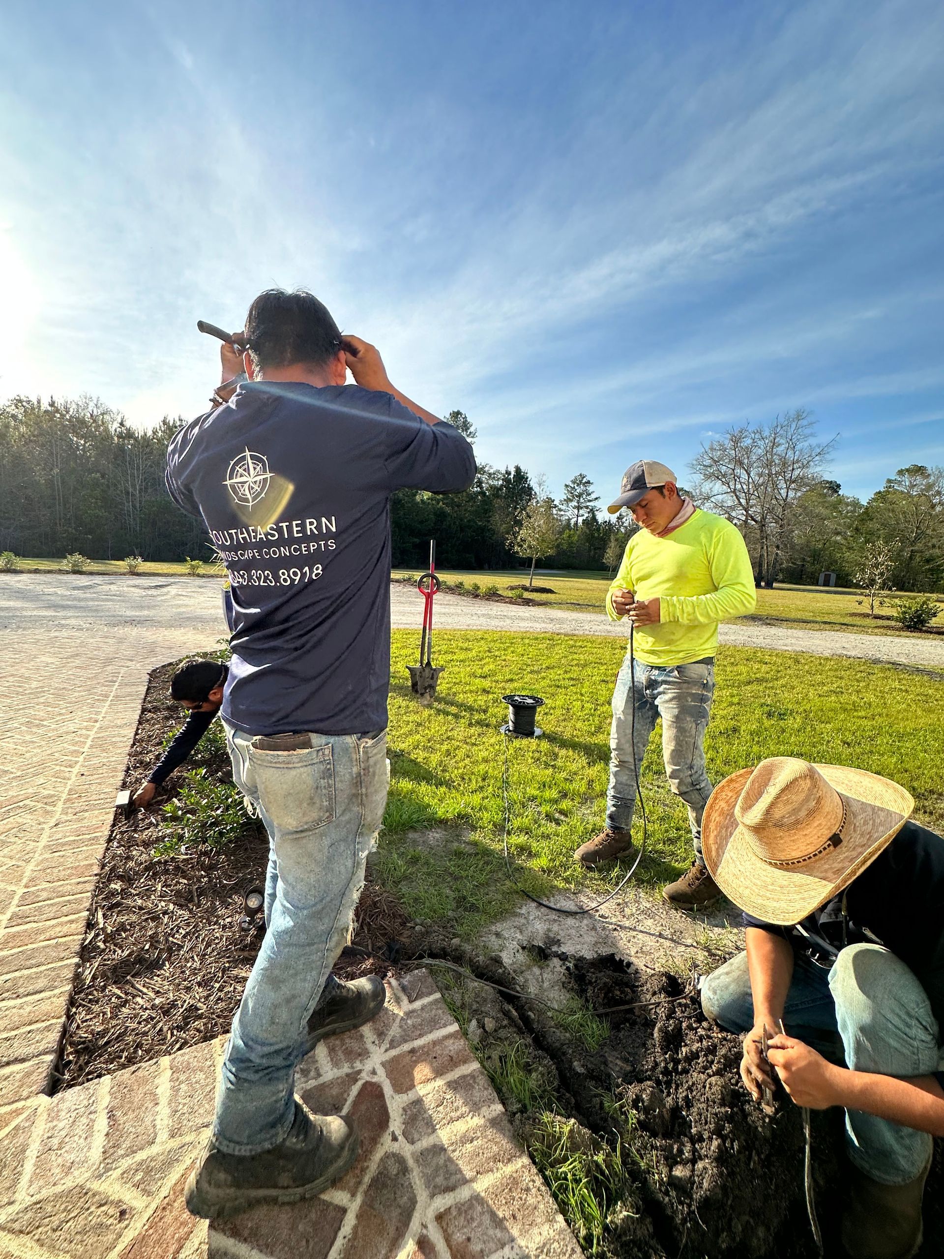 Three workers outdoors; one in blue shirt, one in yellow, one in straw hat. They are working near a brick border.