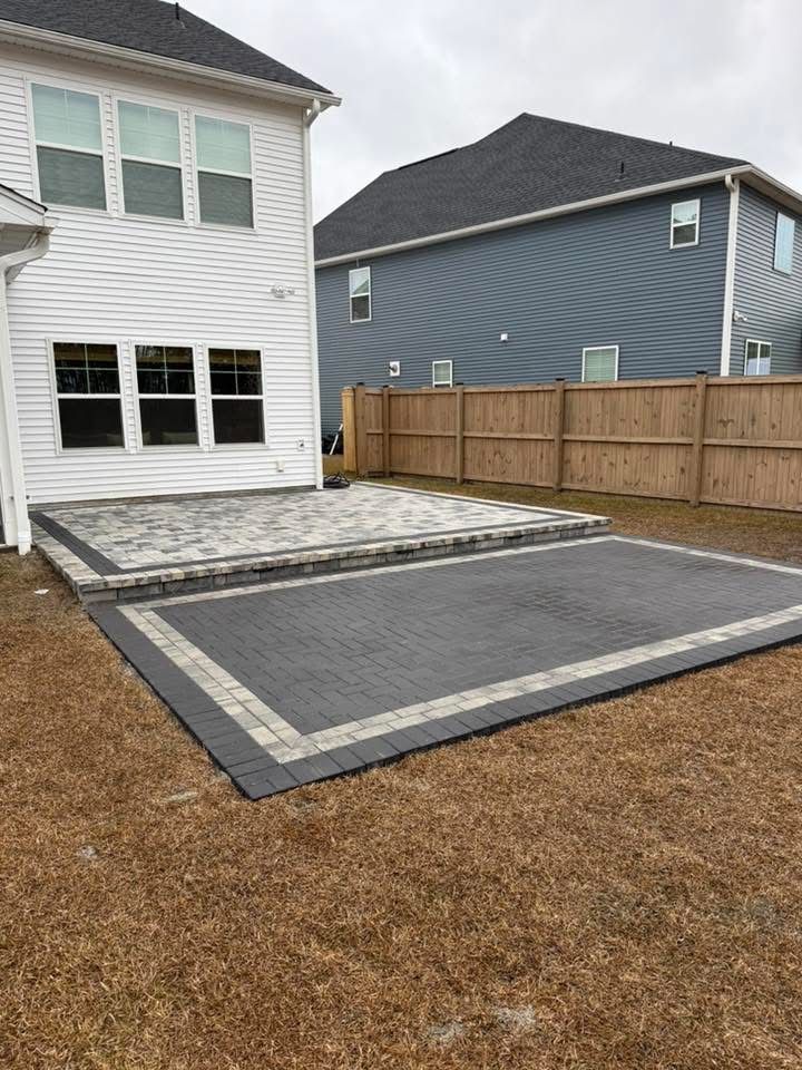 Backyard patio with two levels of pavers. The ground is dry grass, a wooden fence is present.