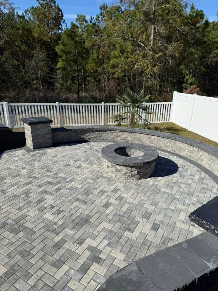 Paver patio with fire pit, bordered by a low wall and white fence, set against a backdrop of trees.