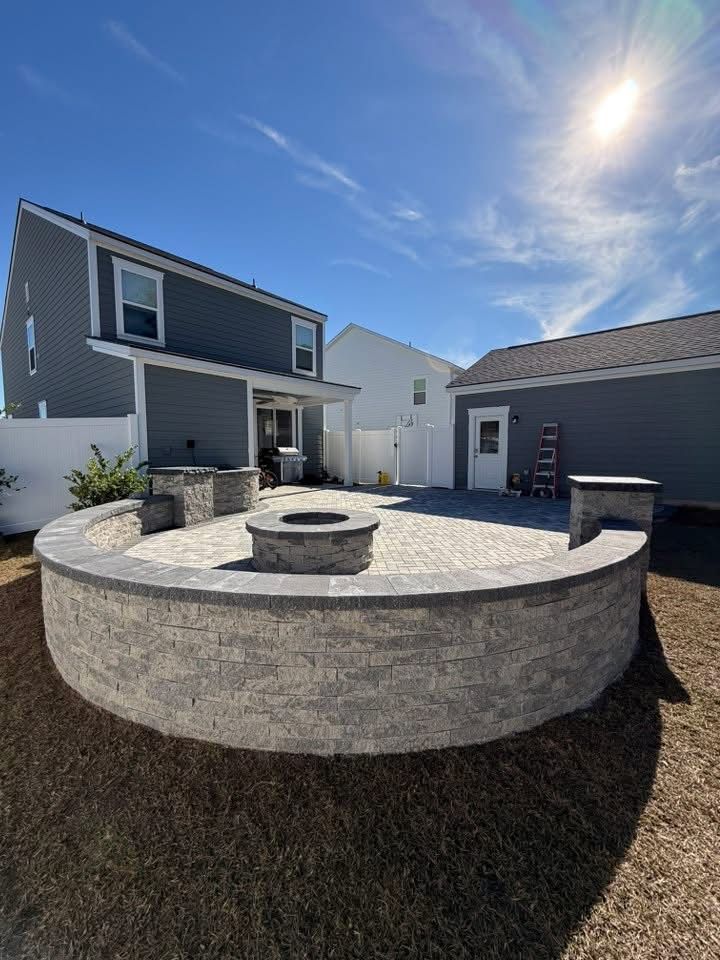 A paver patio with a fire pit, surrounded by a low gray retaining wall, under a blue sky.