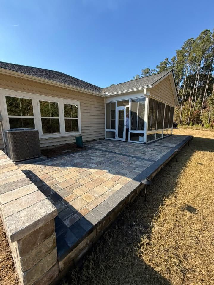 Patio with brick pavers, raised border, and screened-in porch next to a house on a sunny day.