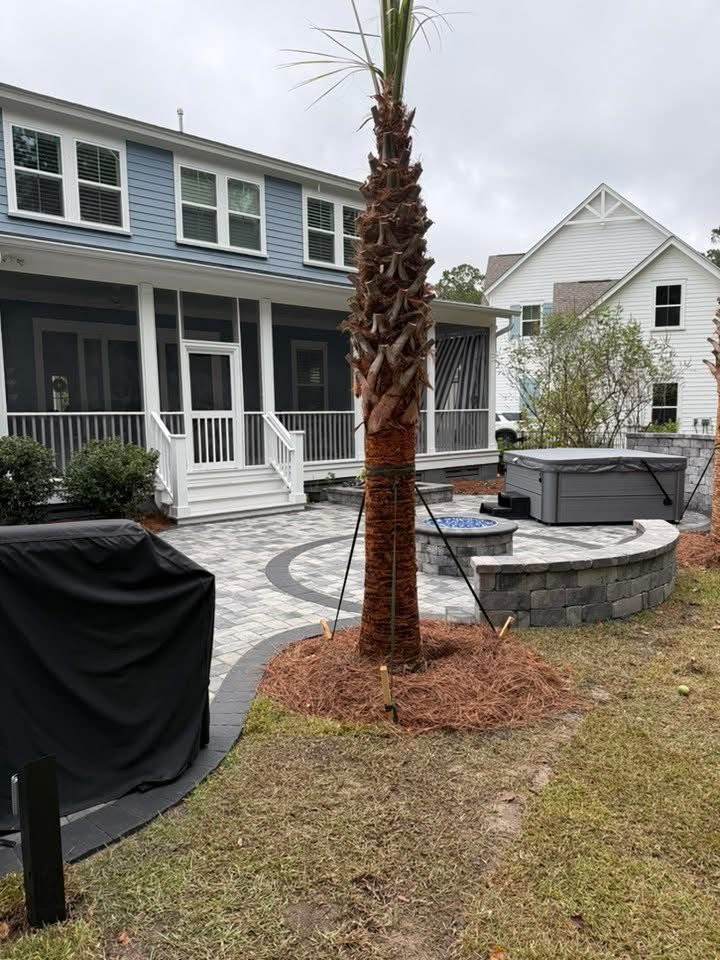 Backyard patio with palm tree, hot tub, and two-story blue house. Brick pavers and a fire pit are also visible.