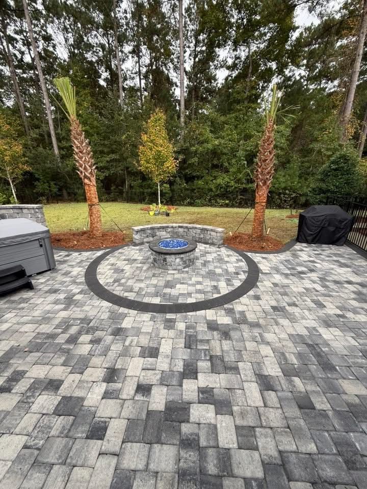 Patio with a fire pit, paved with gray and black bricks, palm trees, and a spa in a backyard setting.