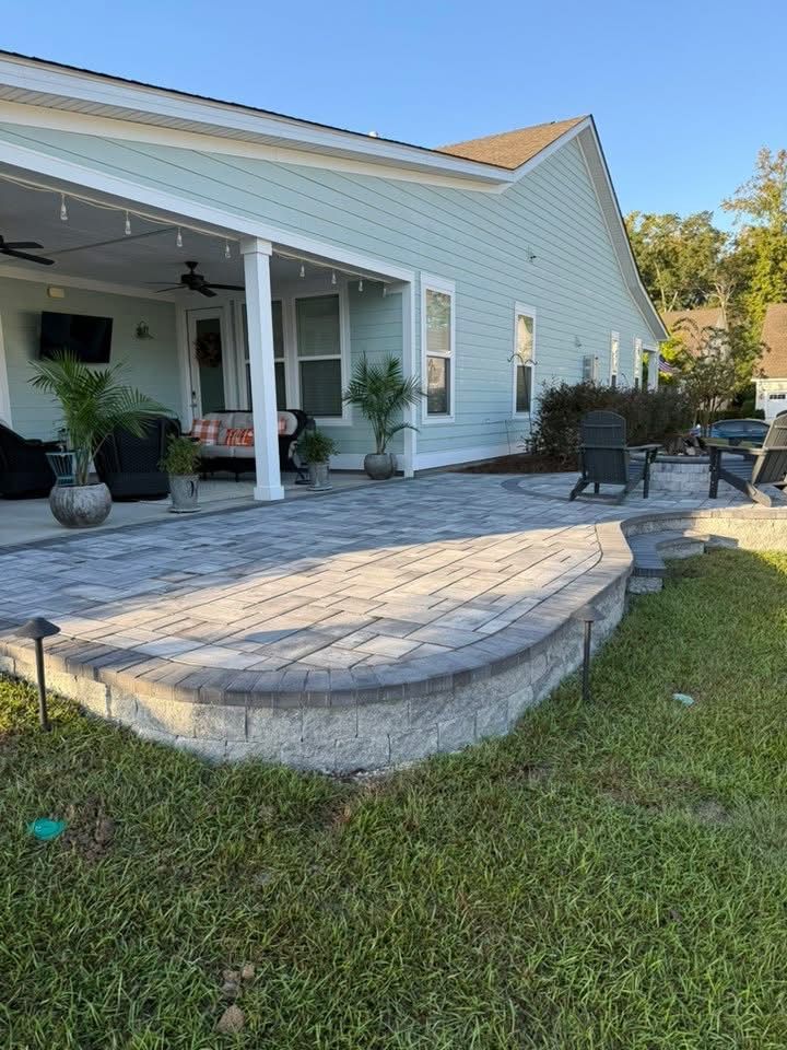 Brick patio with curved retaining wall in front of a light blue house. Outdoor seating on patio under a covered porch.