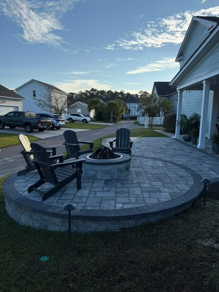Circular patio with fire pit and Adirondack chairs. Suburban neighborhood with houses and cars.