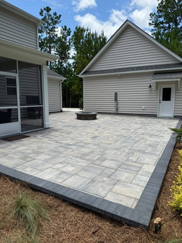 Stone patio with fire pit, bordered by dark pavers and landscaping, adjacent to a house with a screened porch and garage.