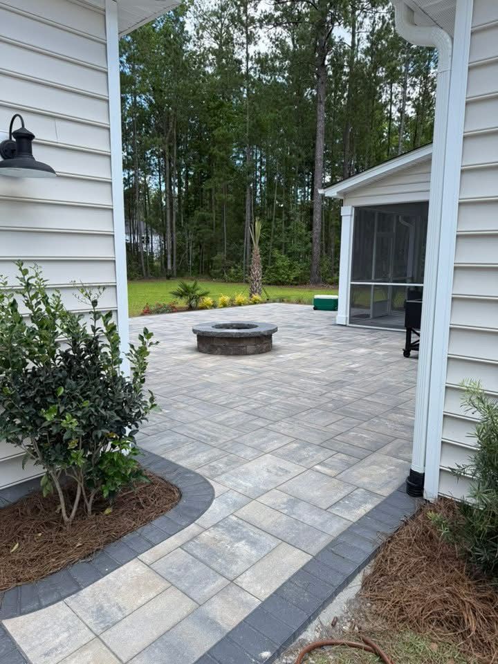 Patio with stone pavers, fire pit, and screened-in porch, framed by a white archway and foliage.