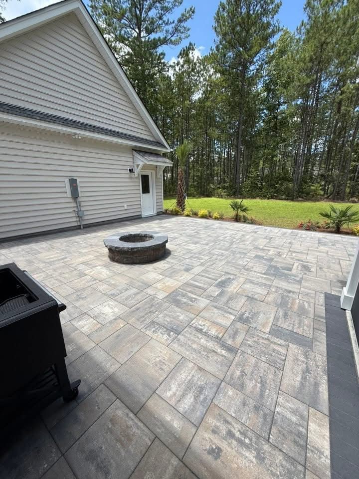 Paver patio with a fire pit in front of a house, surrounded by grass and trees.