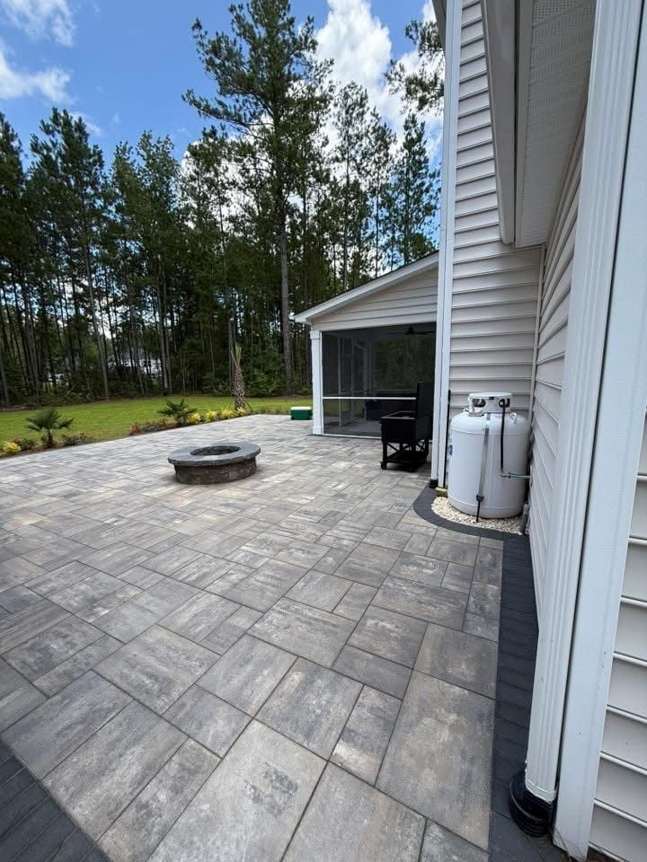 Stone patio with fire pit, grill, and propane tank next to a white house with trees in the background.