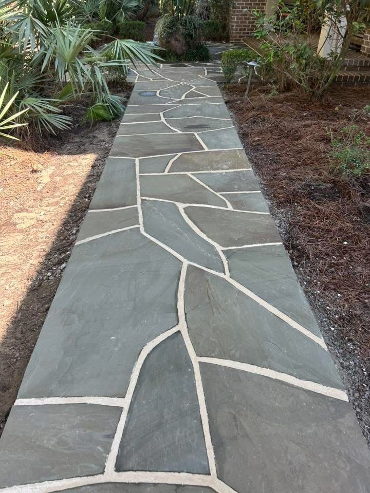 Stone pathway with irregular shapes and light-colored grout, bordered by plants and dirt.