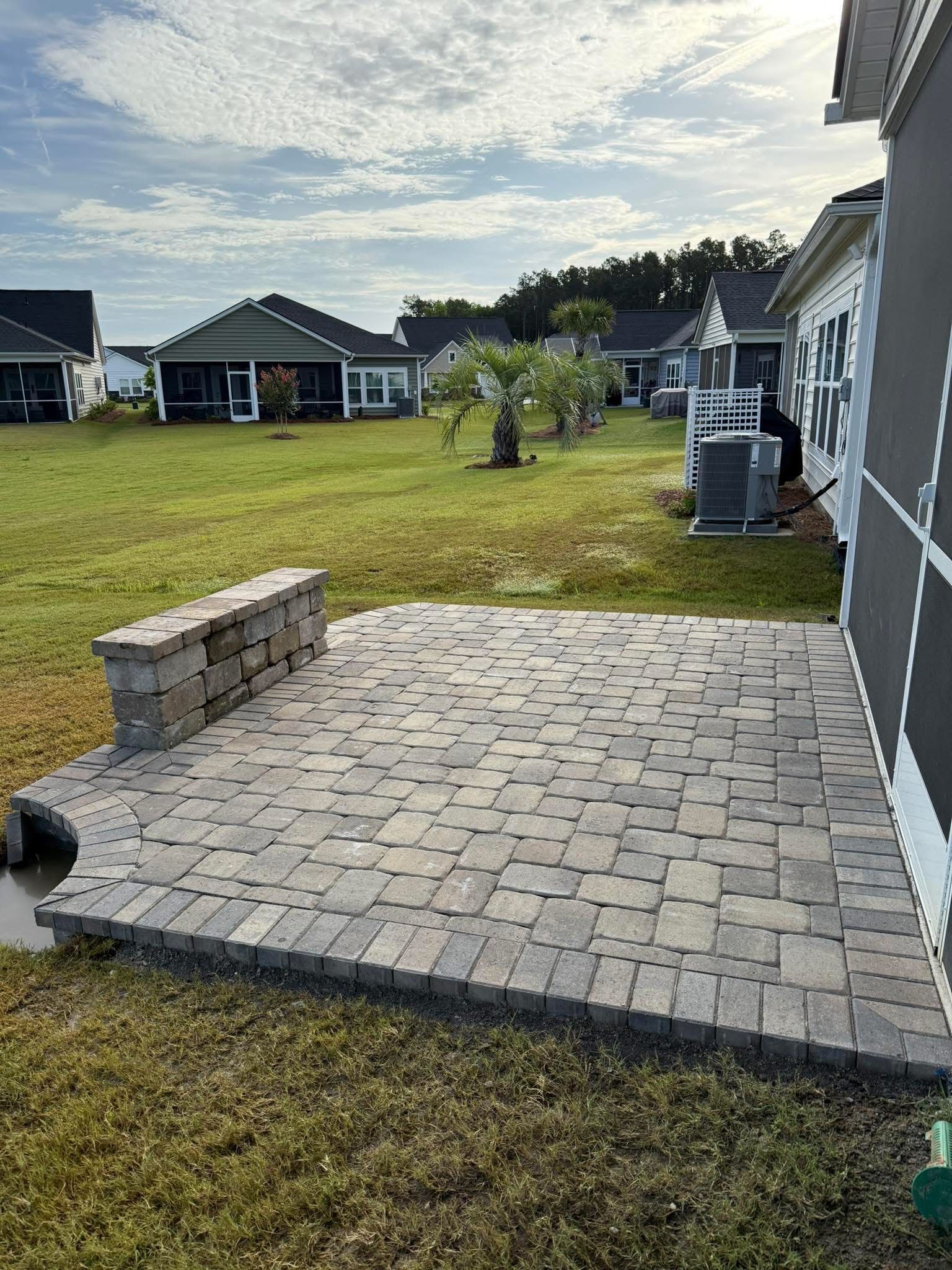 Brick patio next to a house with a low retaining wall. Green grass and other houses in the background.