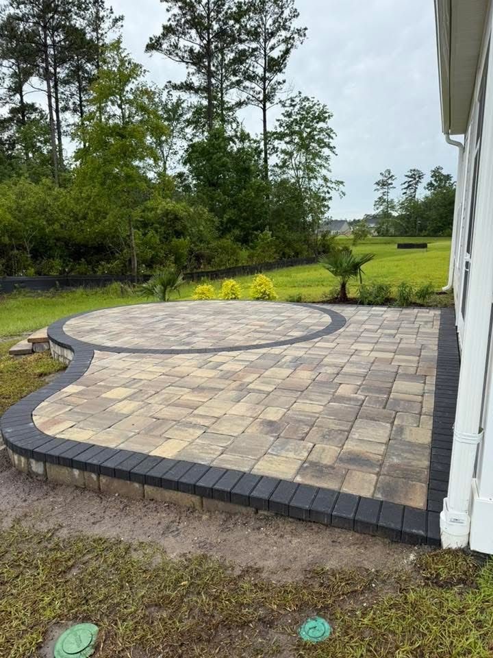 Brick patio with dark border next to a white house, grass, and trees.