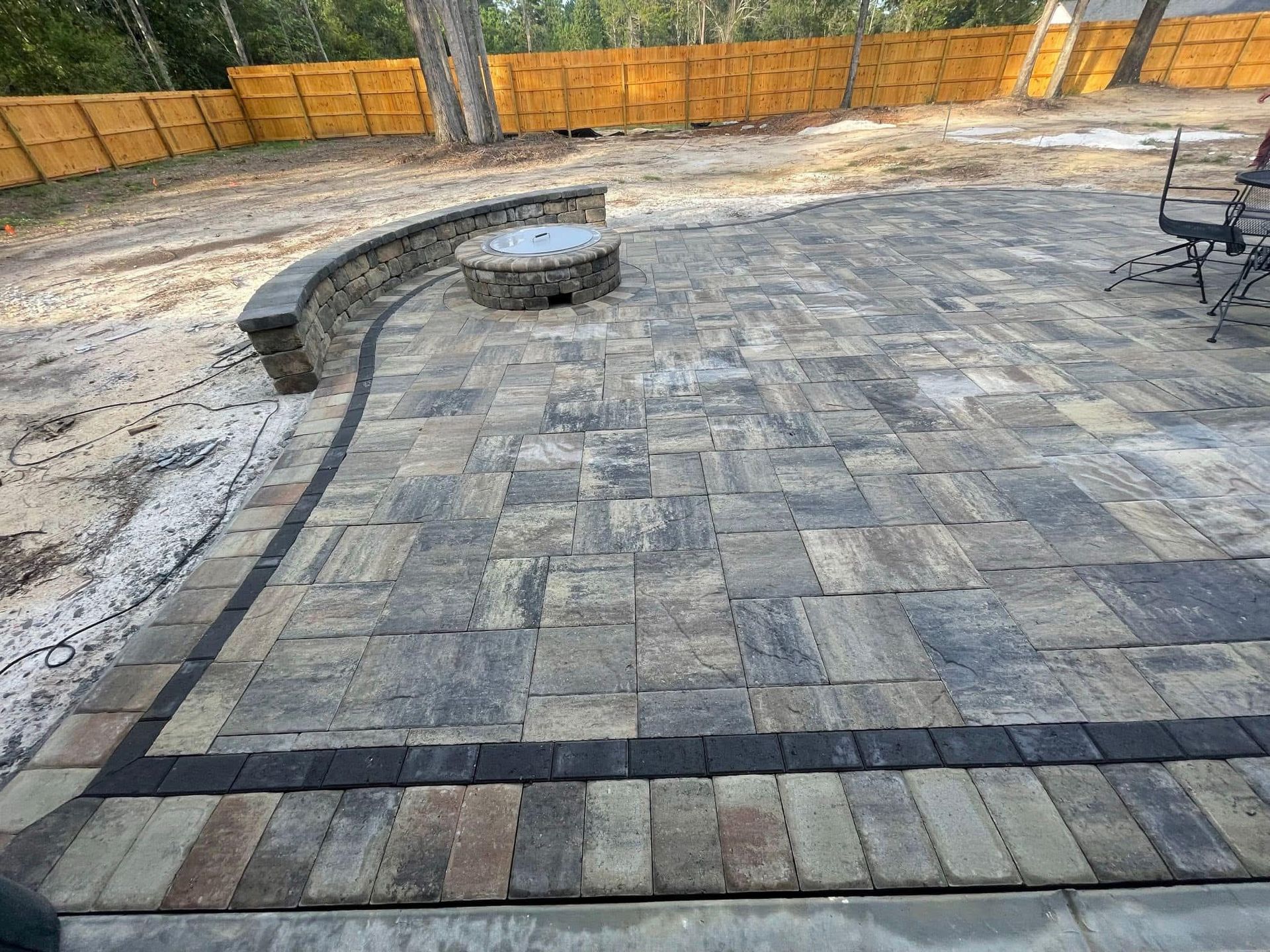 Brick patio with fire pit and curved seating wall, bordered by dark bricks, against a wooden fence.