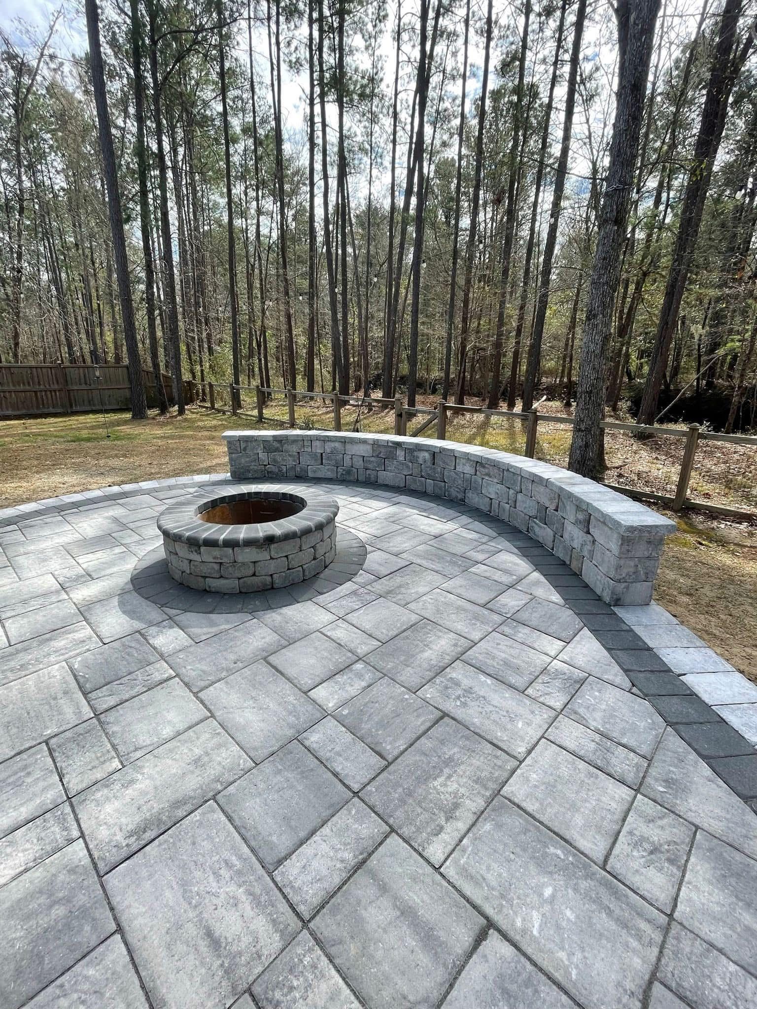 Patio with fire pit and curved stone wall in a wooded area. Gray pavers and trees in the background.
