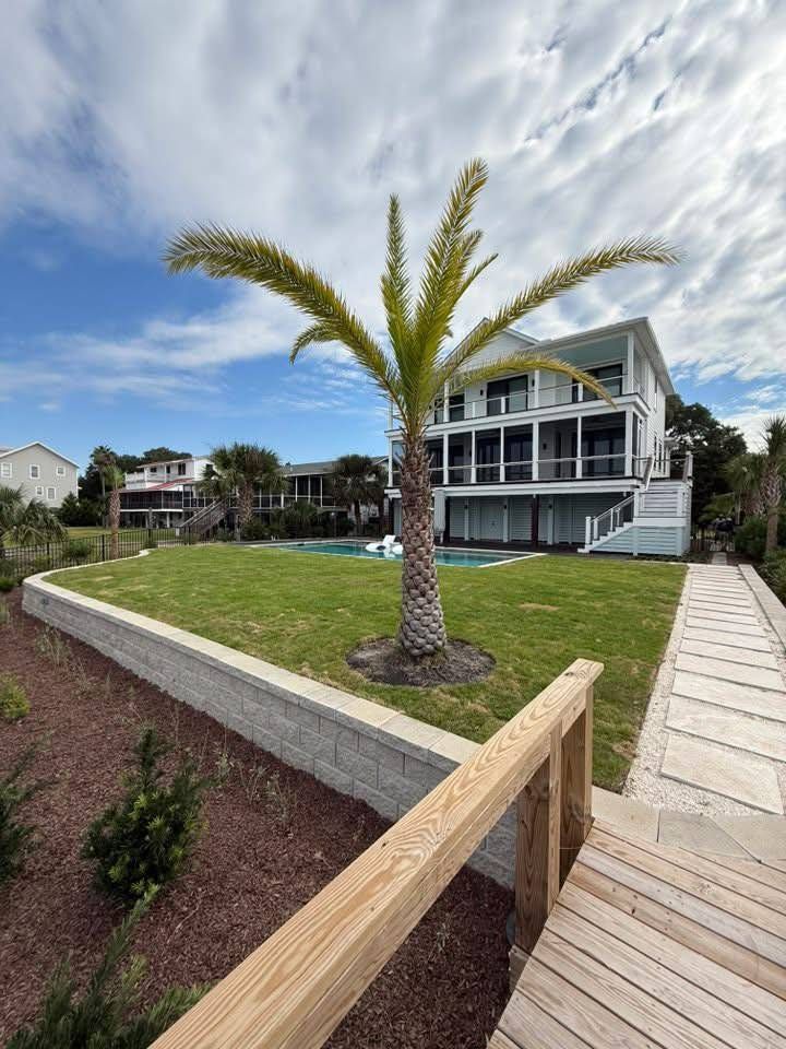 Modern white beach house with palm tree, green lawn, pool, and wooden walkway against a blue sky.
