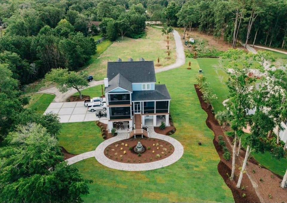 Two-story blue house with porch, circular flower bed, green lawn, trees and gravel driveway.