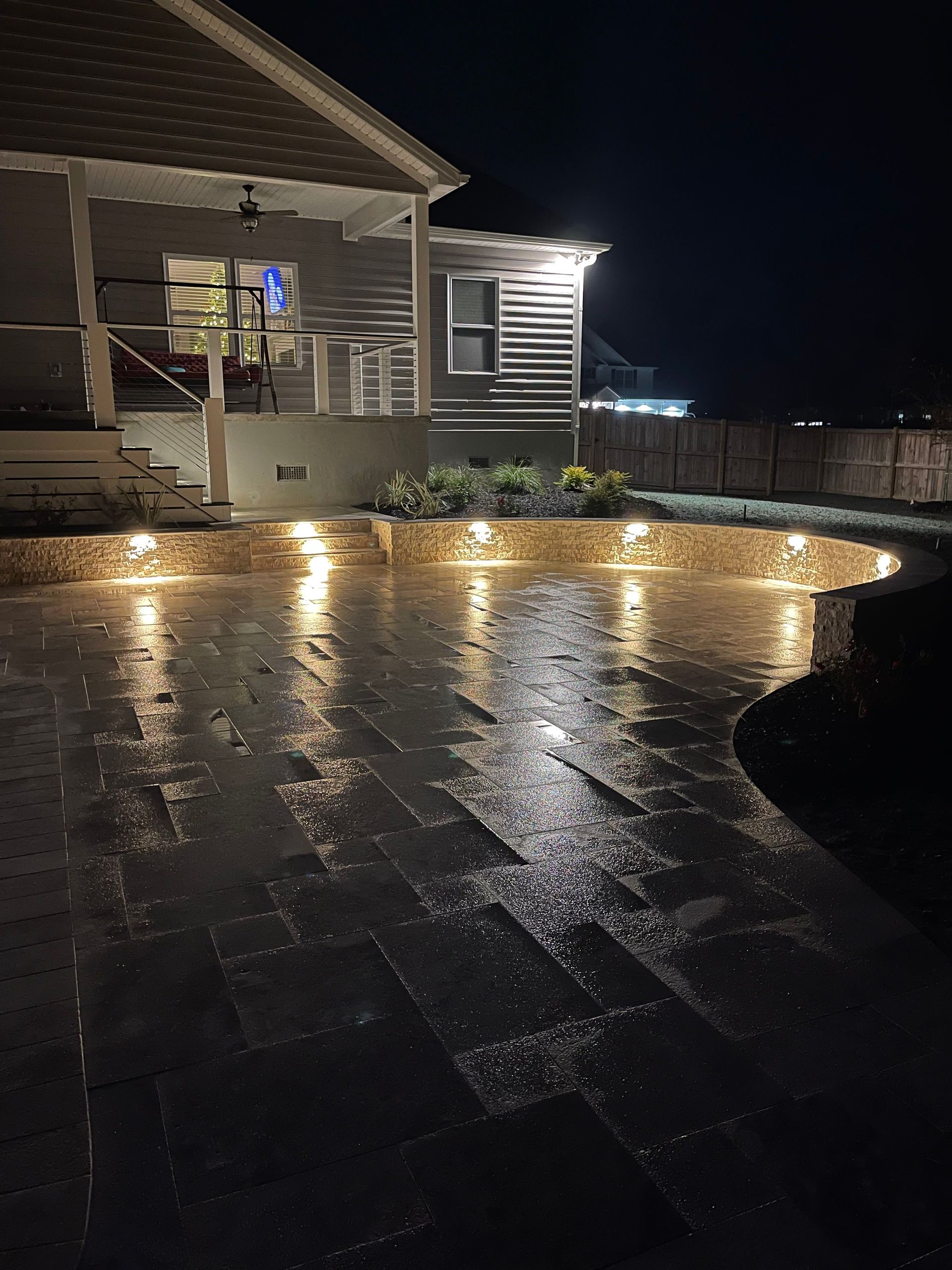 Brick patio illuminated at night with lights, reflecting wet surface. Building in background.