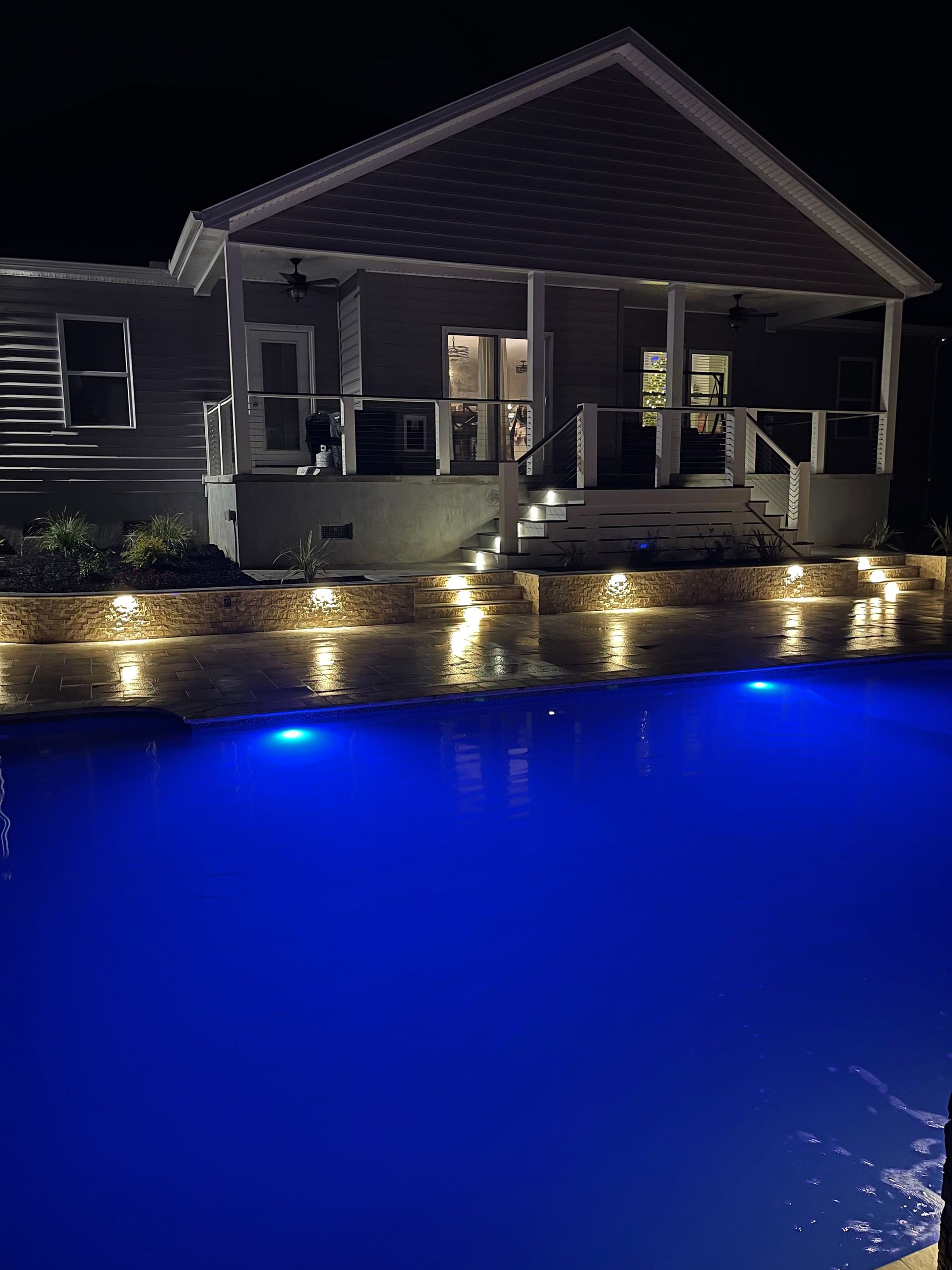 Nighttime view of a house with a pool lit with blue lights. Warm lights illuminate the patio and landscaping.