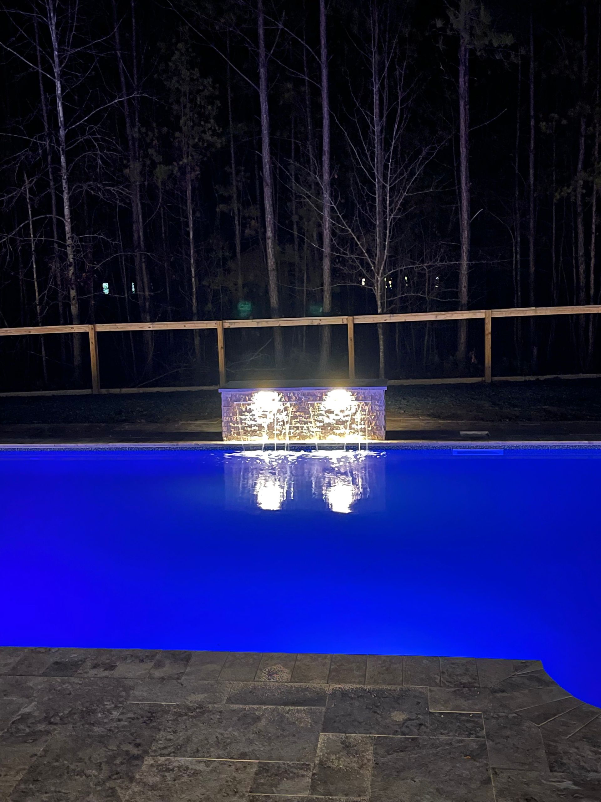 Nighttime pool with blue water and illuminated waterfall feature, trees in the background.