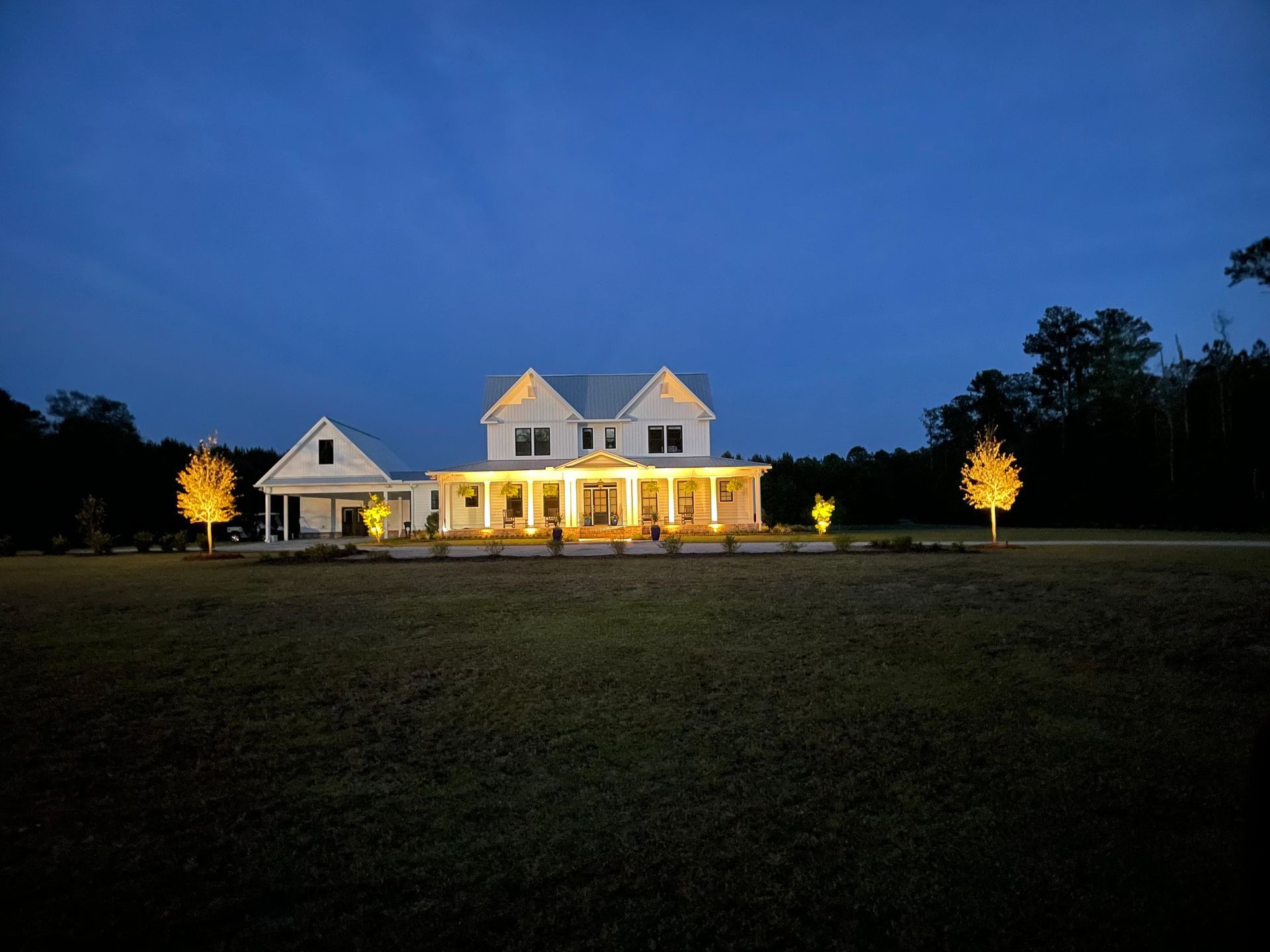 White farmhouse with porch illuminated at dusk. Two trees on either side are also lit.