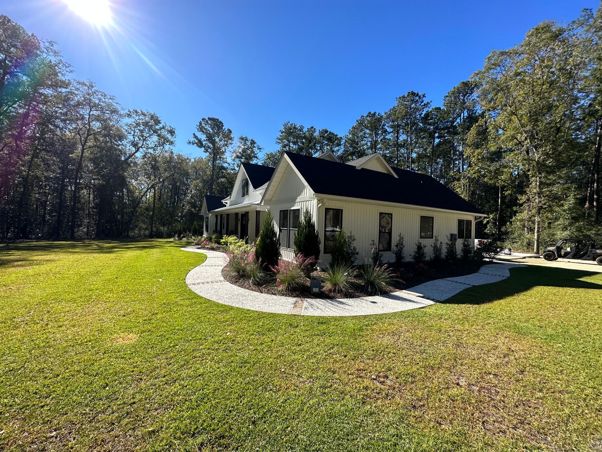White house with black roof, pathway, surrounded by green grass and trees under a clear blue sky.