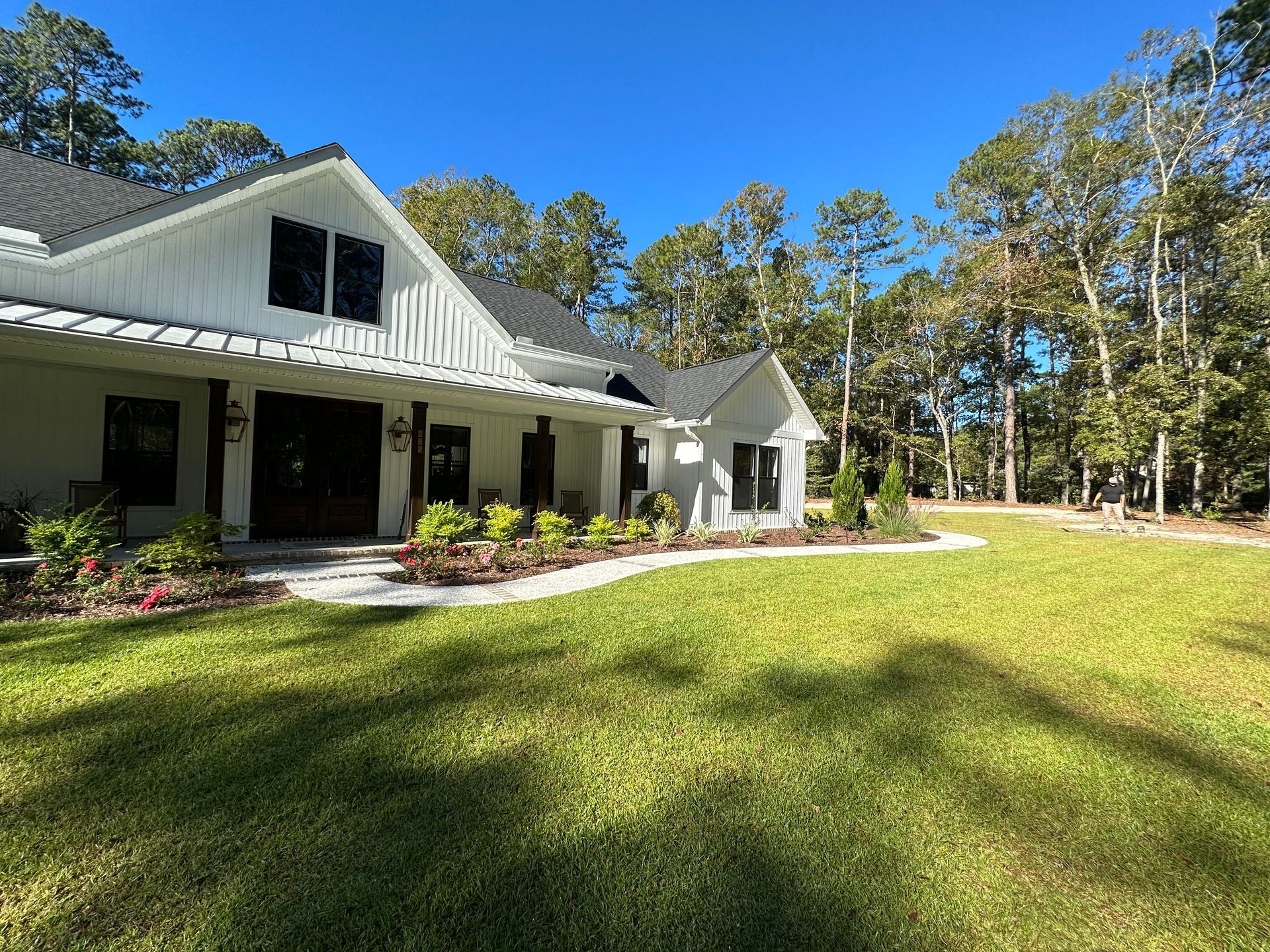 White farmhouse with black trim and roof, on a green lawn, surrounded by trees under a blue sky.