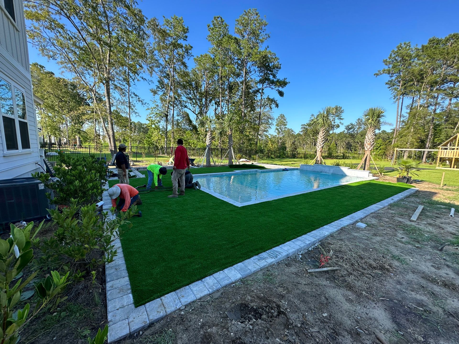 Workers installing artificial turf around a pool with blue water. Bright green grass, gray pavers, sunny outdoor setting.