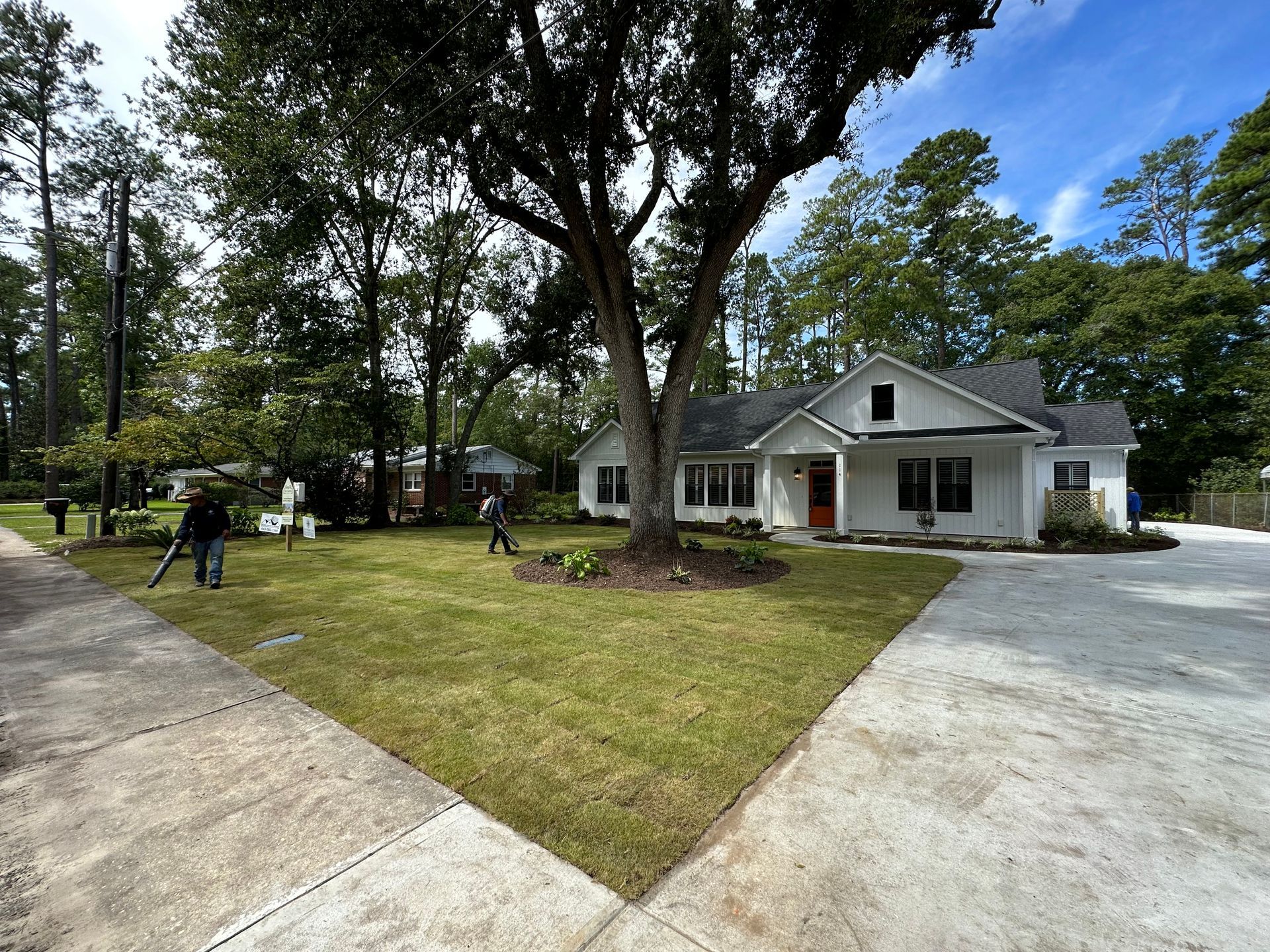 White house with red door, large tree, and green lawn; people tending the yard.