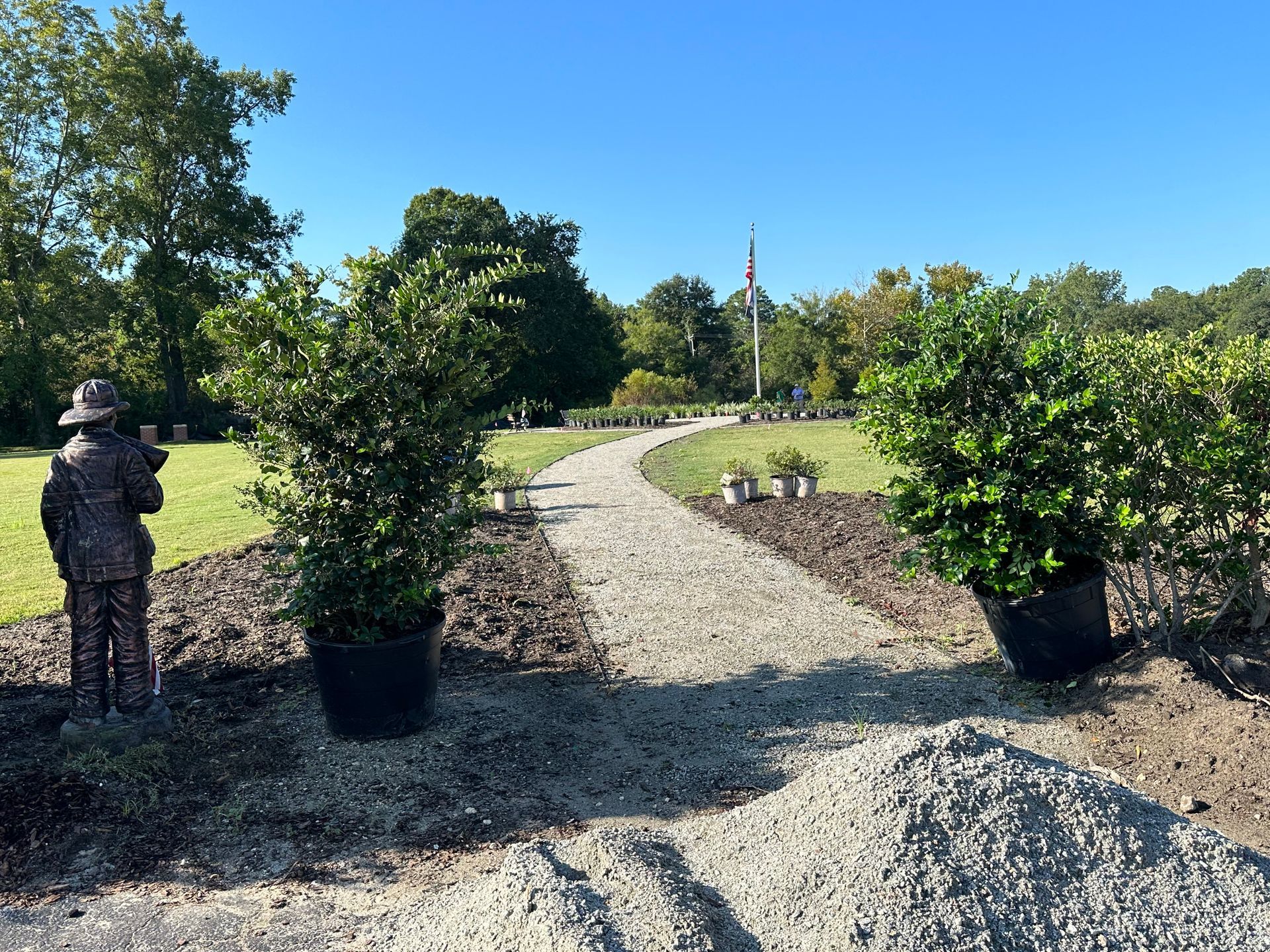 Gravel path curves through a garden, lined with bushes, leading to a flagpole under a blue sky. A statue is on the left.