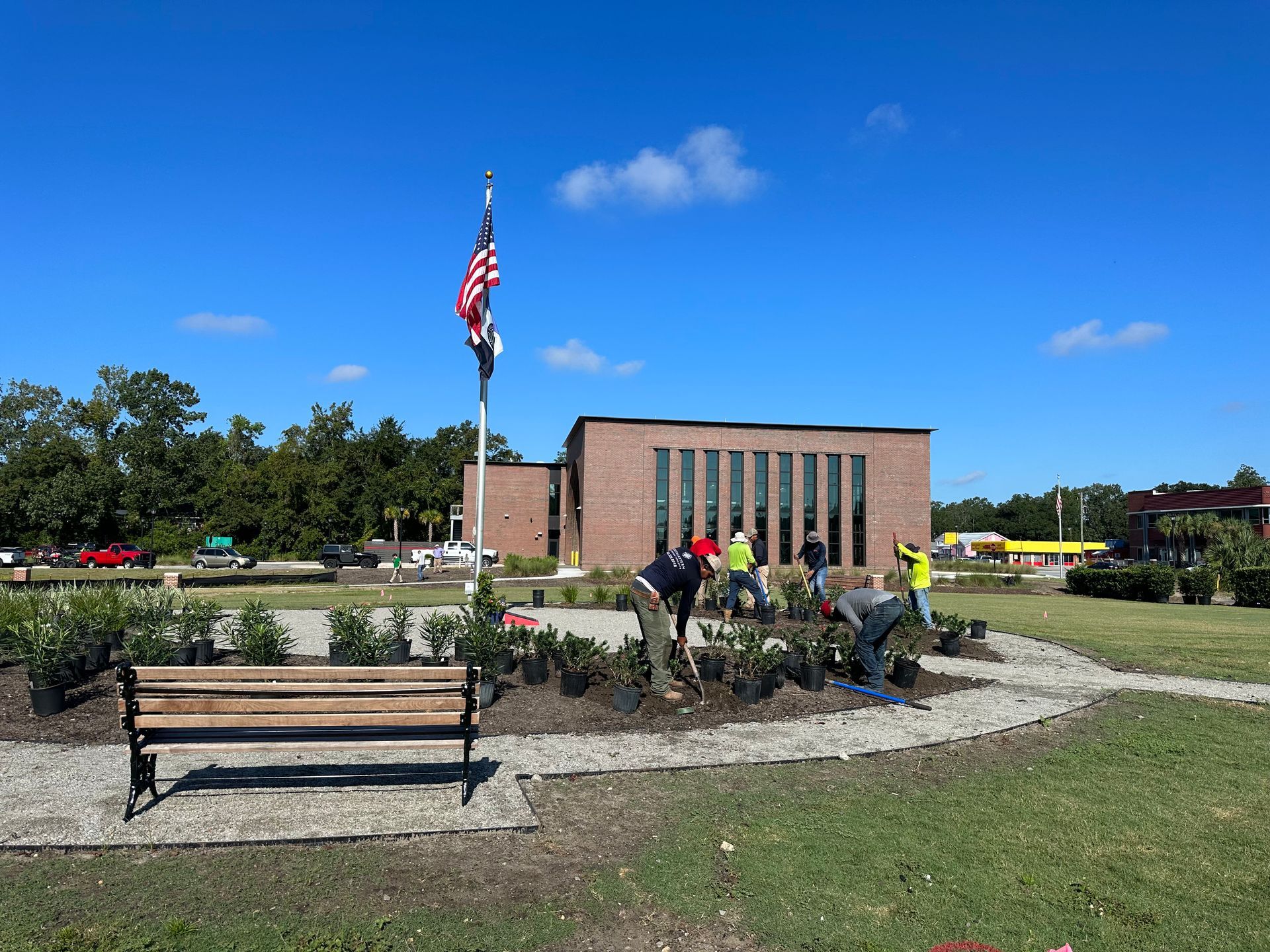 People planting in a garden with a flag and a brick building in the background. A bench is in the foreground.
