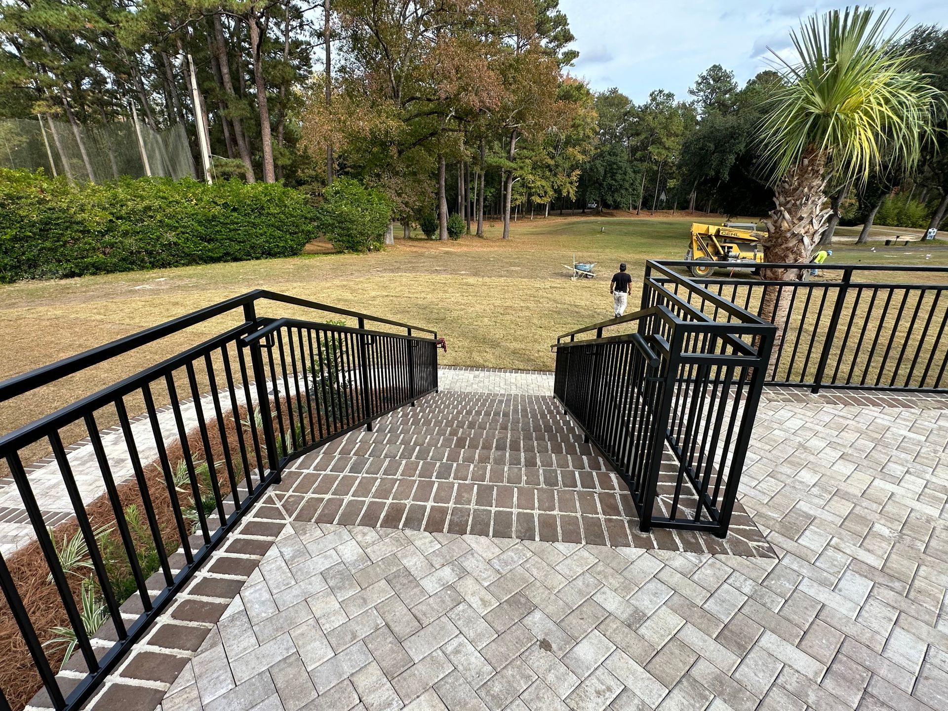 Black railing leads down brick steps to a grassy yard. A person walks in the distance.