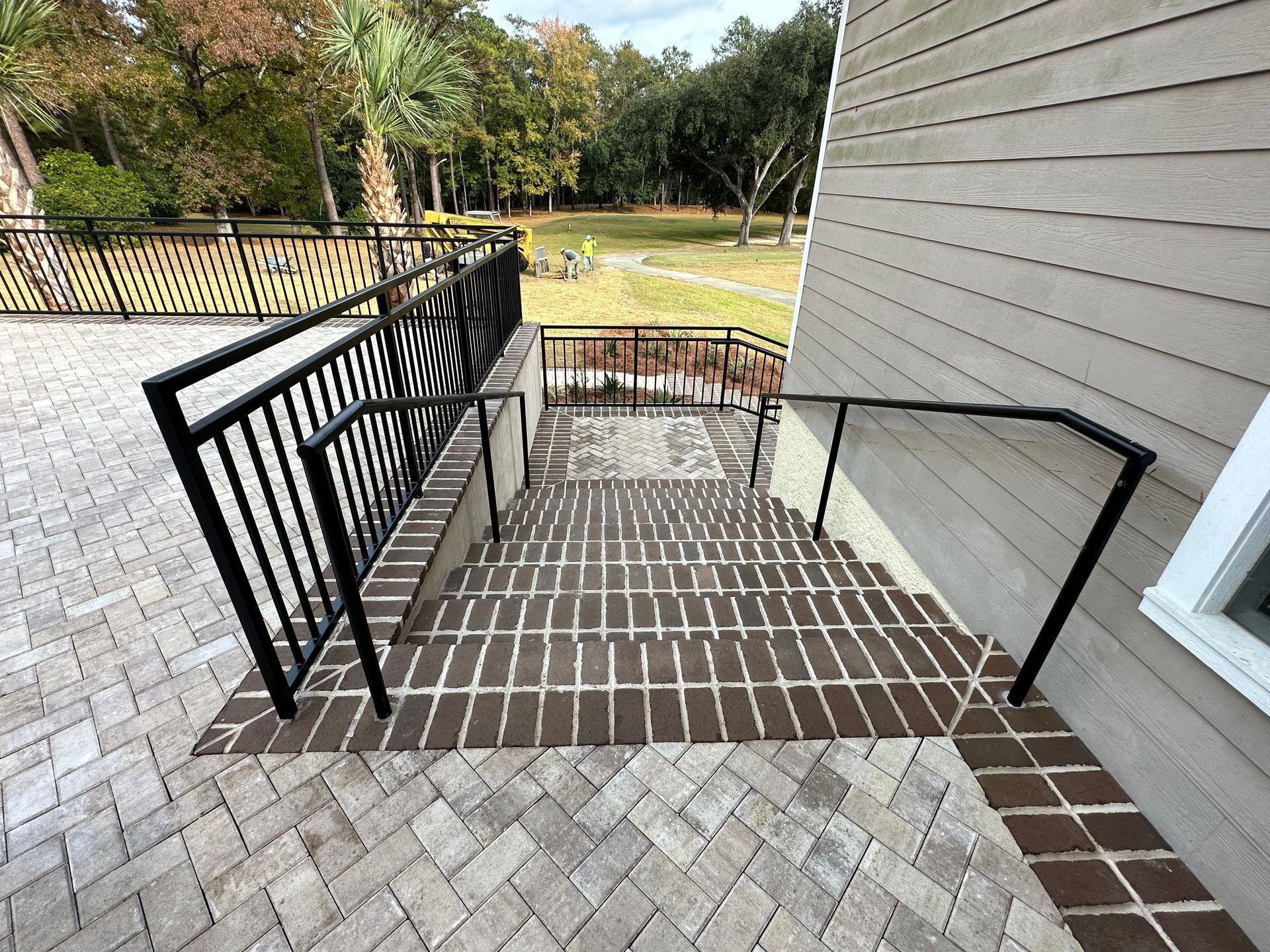 Black metal handrails on brick steps leading to a paved patio, beside a tan building.