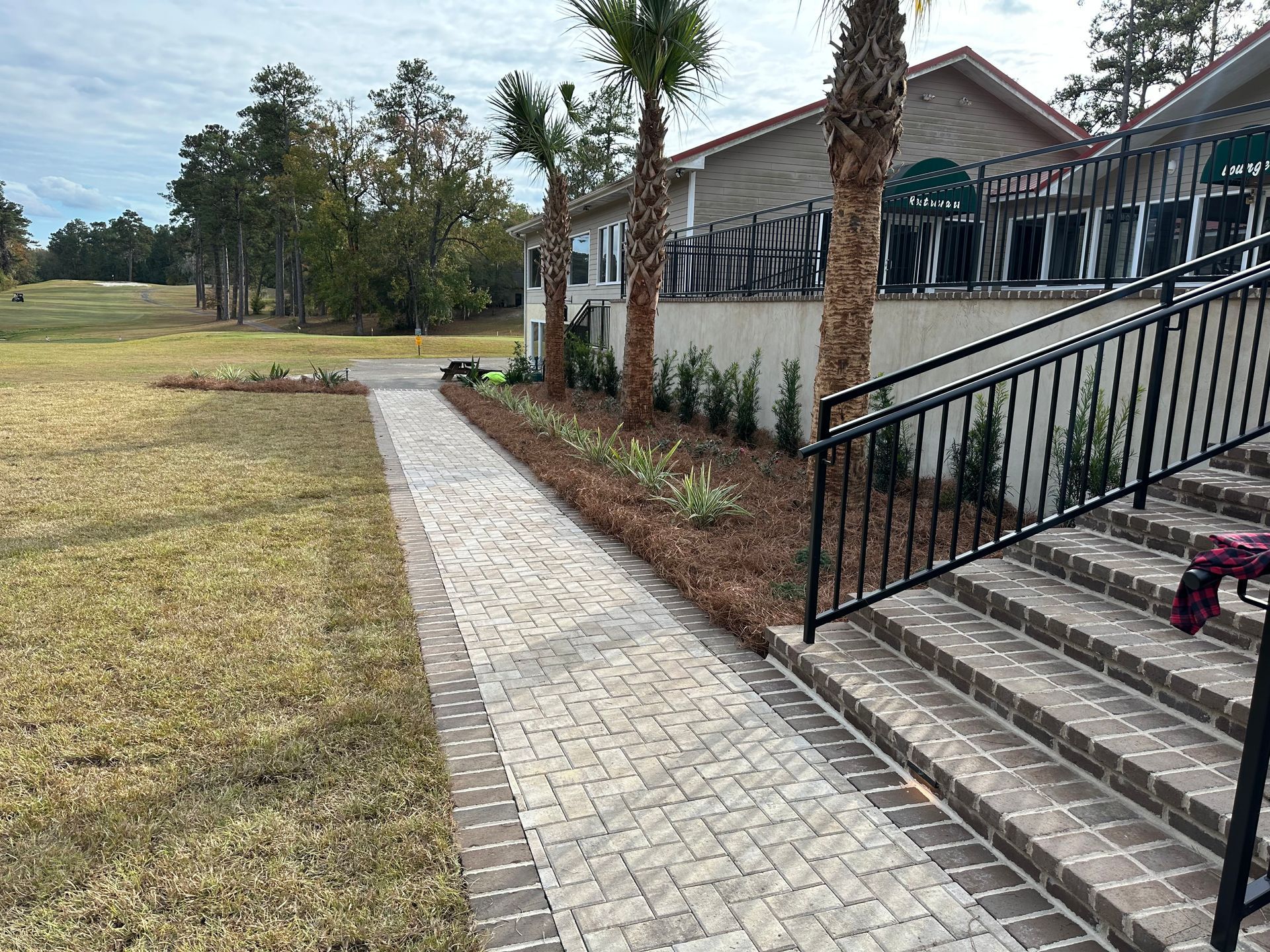 Brick pathway leading to a building with steps and a black railing, next to a grassy lawn and a golf course.
