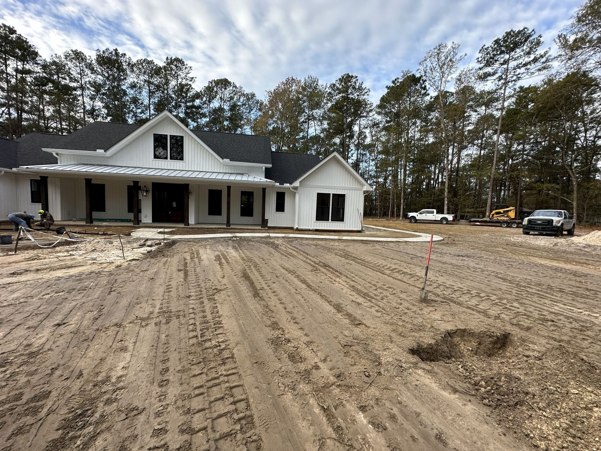 White farmhouse under construction, dirt lot, two trucks, and trees under a cloudy sky.