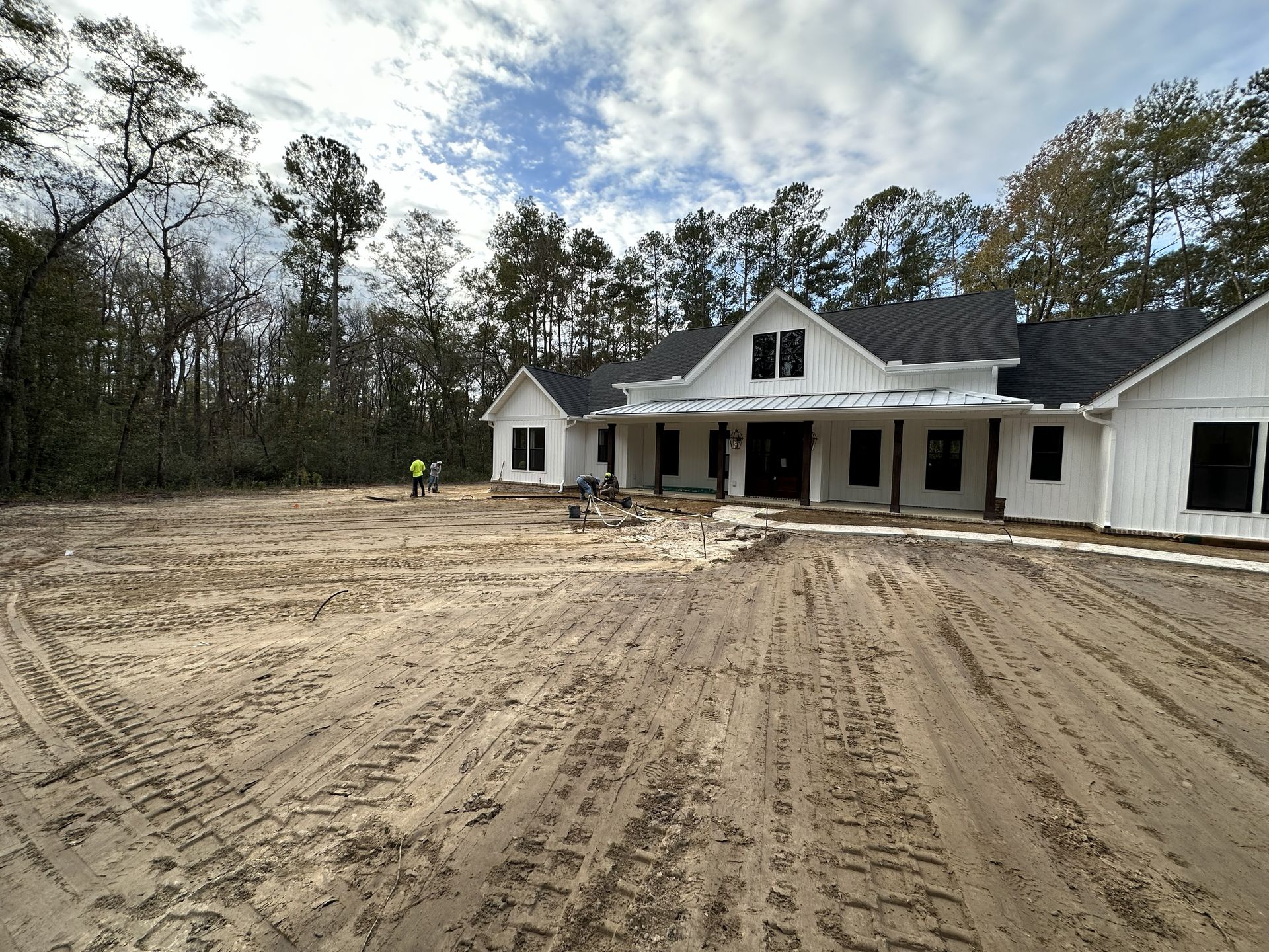 White farmhouse under construction on a dirt lot, with dark roof and trim, surrounded by trees.