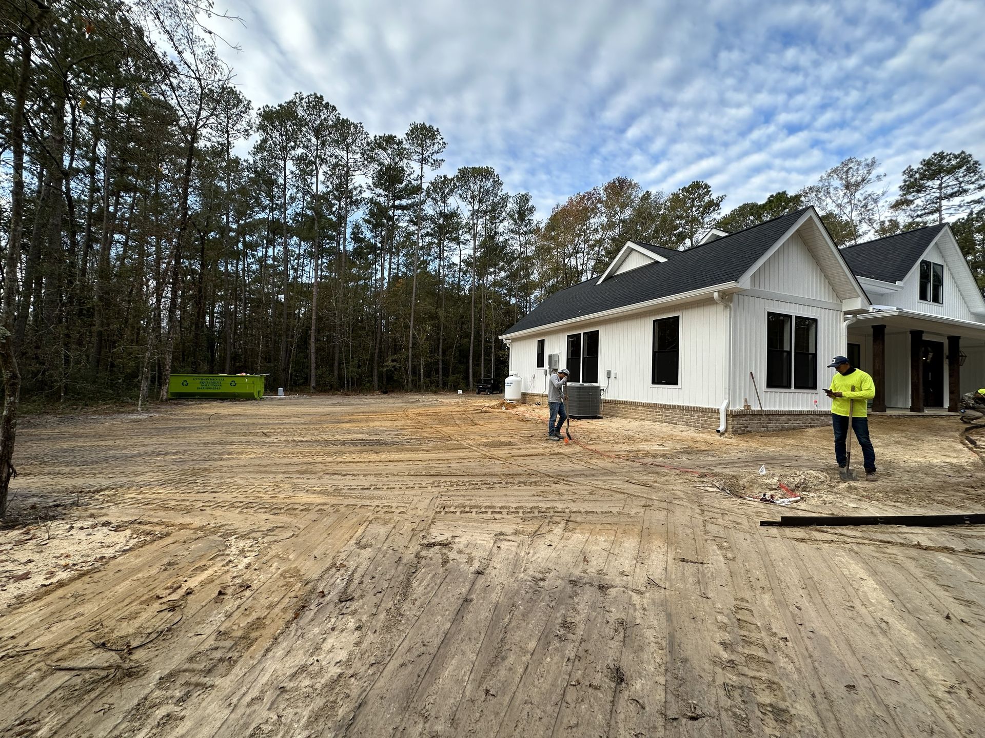 Construction site with new white house. Two workers near house, dirt foreground, trees and sky background.