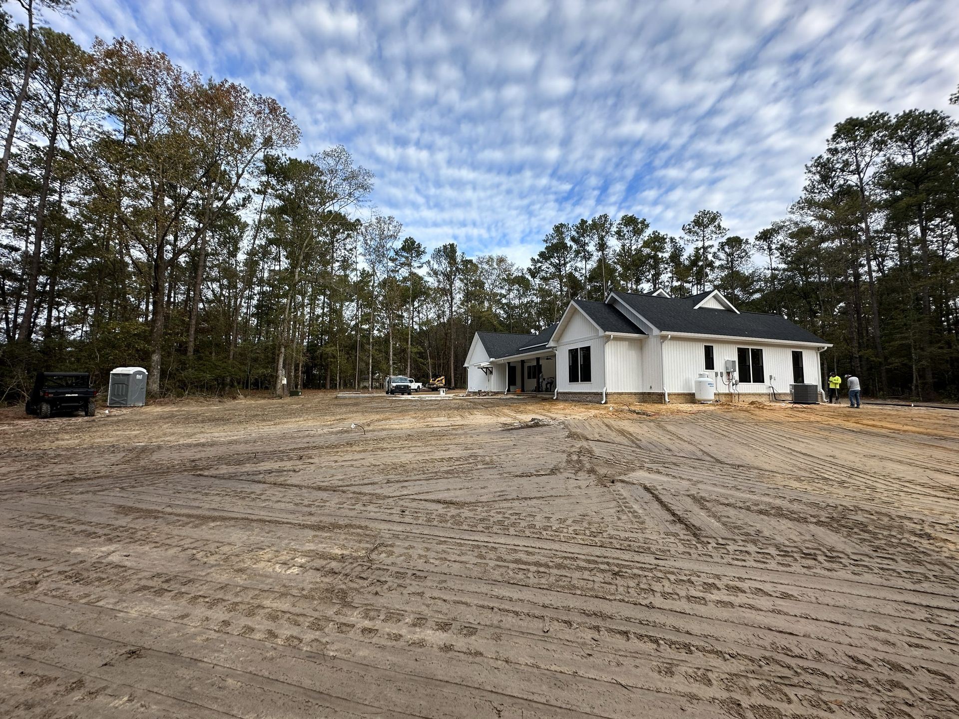 A new white house under construction in a sandy lot surrounded by trees, cloudy sky overhead.