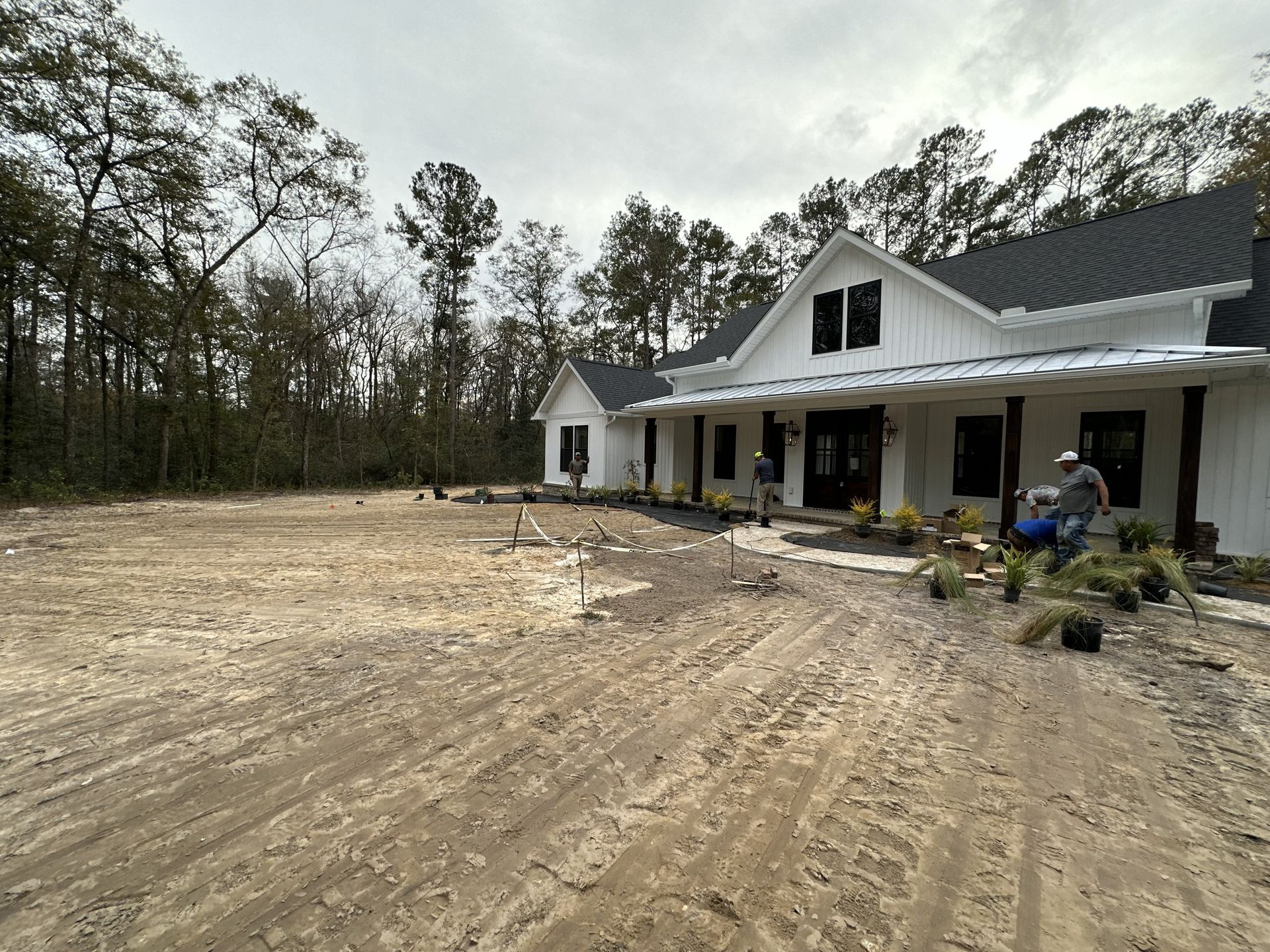 Farmhouse under construction, sandy yard, workers, cloudy day, landscaping underway.