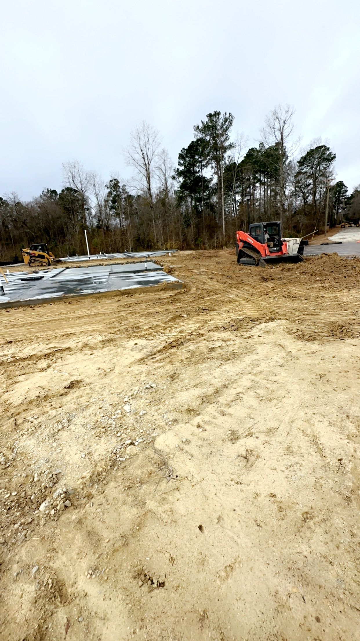 Construction site with a skid steer, muddy ground, and trees under a cloudy sky.