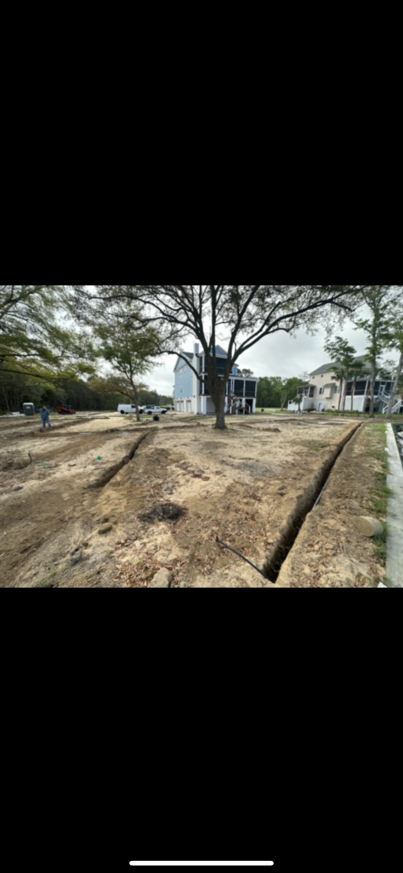 Trenches dug in a dirt yard, with a tree and houses in the background. Cloudy sky.