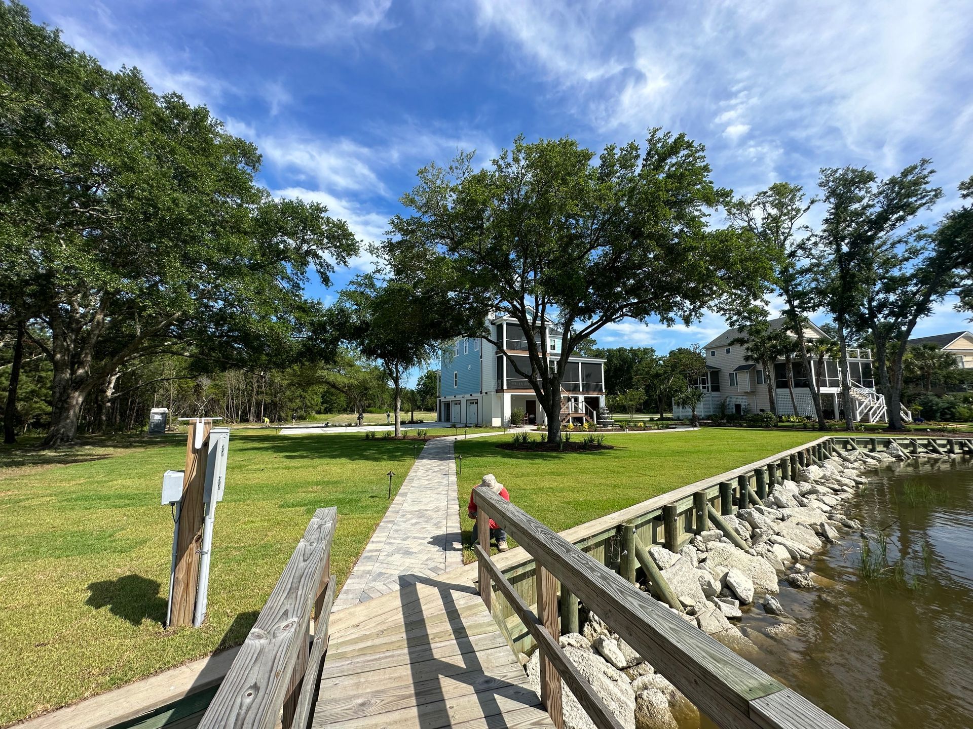 Wooden dock extending into water with a grassy yard and houses under a partly cloudy sky.